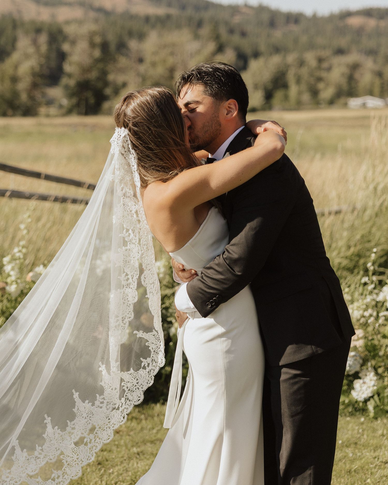 A romantic wedding kiss between newlyweds in an outdoor field setting with a flowing lace veil.