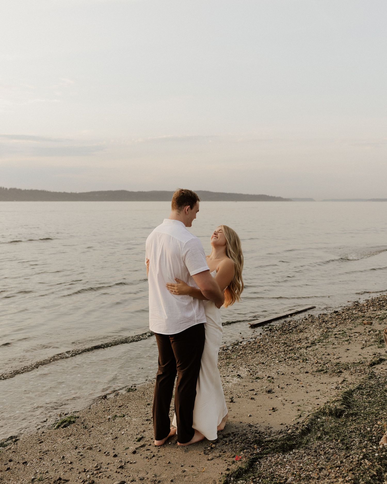 Couple in white and black attire embracing on rocky beach at sunset.