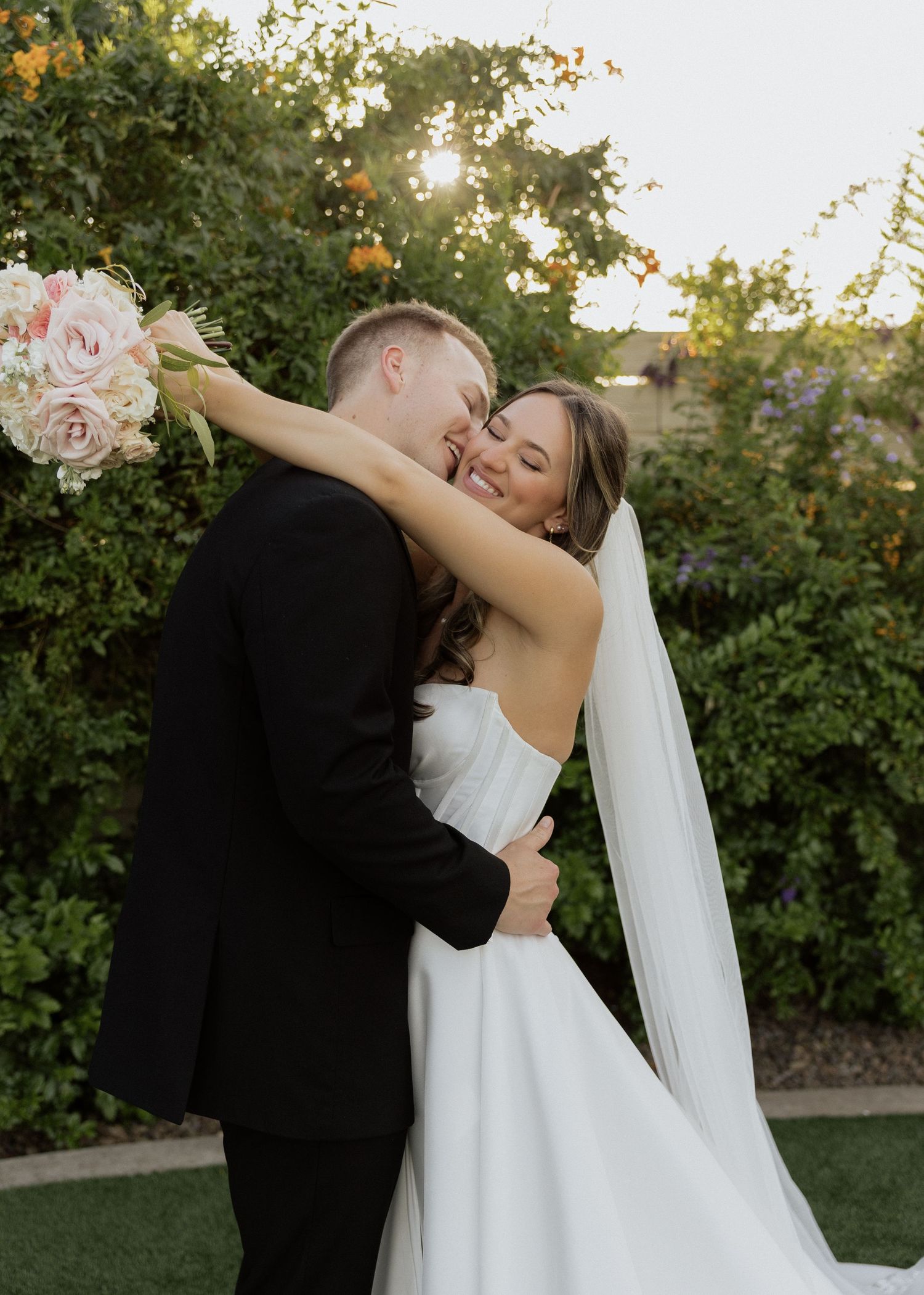 Bride and groom embracing outdoors, bride holding pink bouquet and wearing white gown with veil, groom in black suit.