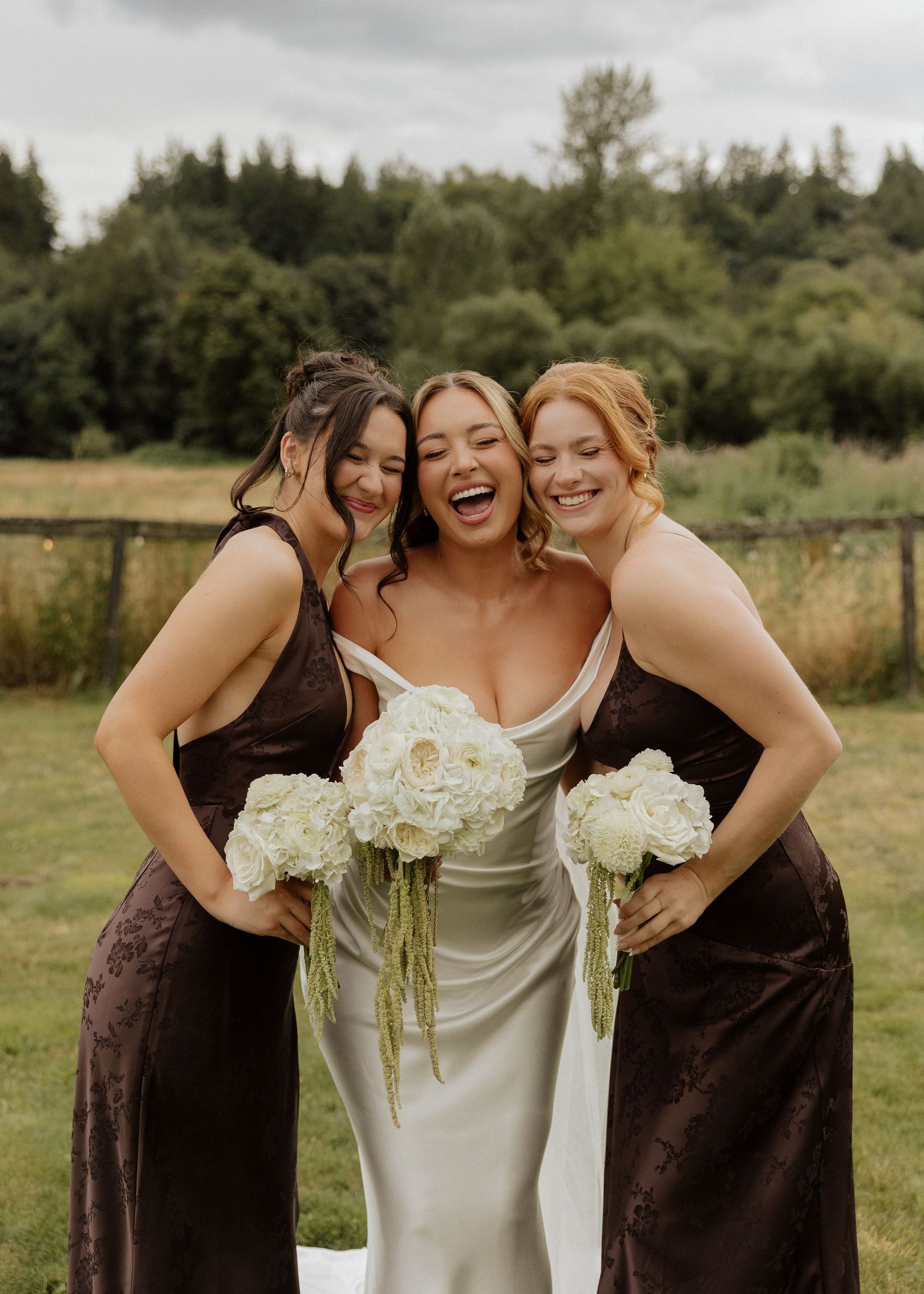 A laughing bride in a white satin gown holds white hydrangeas flanked by bridesmaids in brown dresses.
