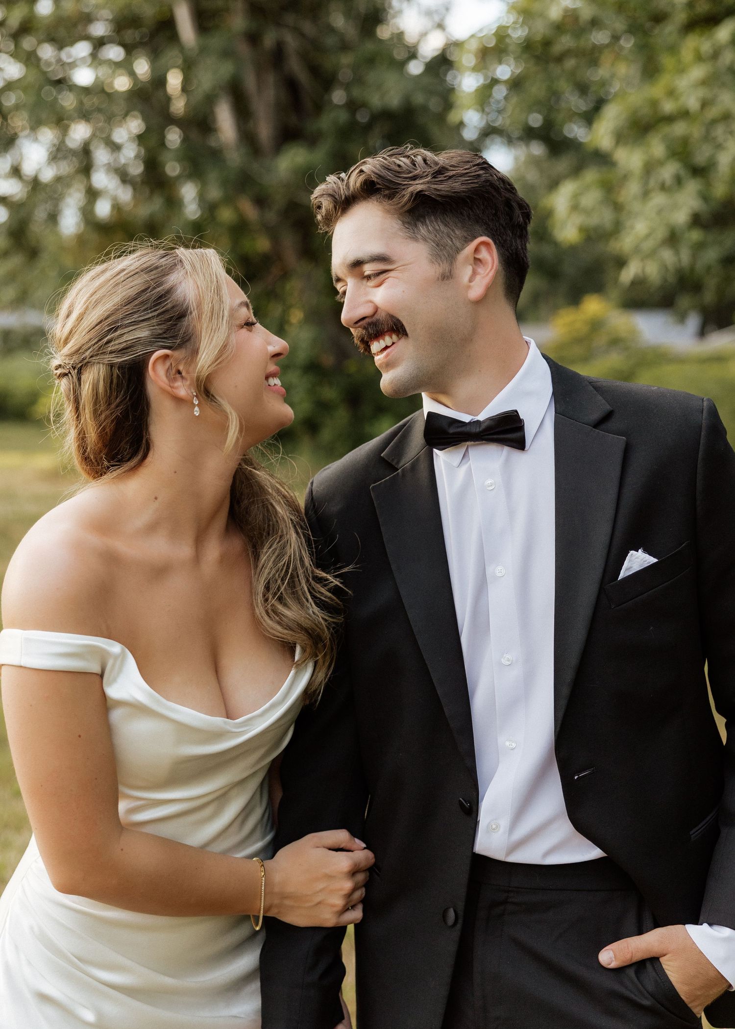 Smiling bride and groom laughing together outdoors, bride in off-shoulder white gown, groom in classic black tuxedo with bow tie.