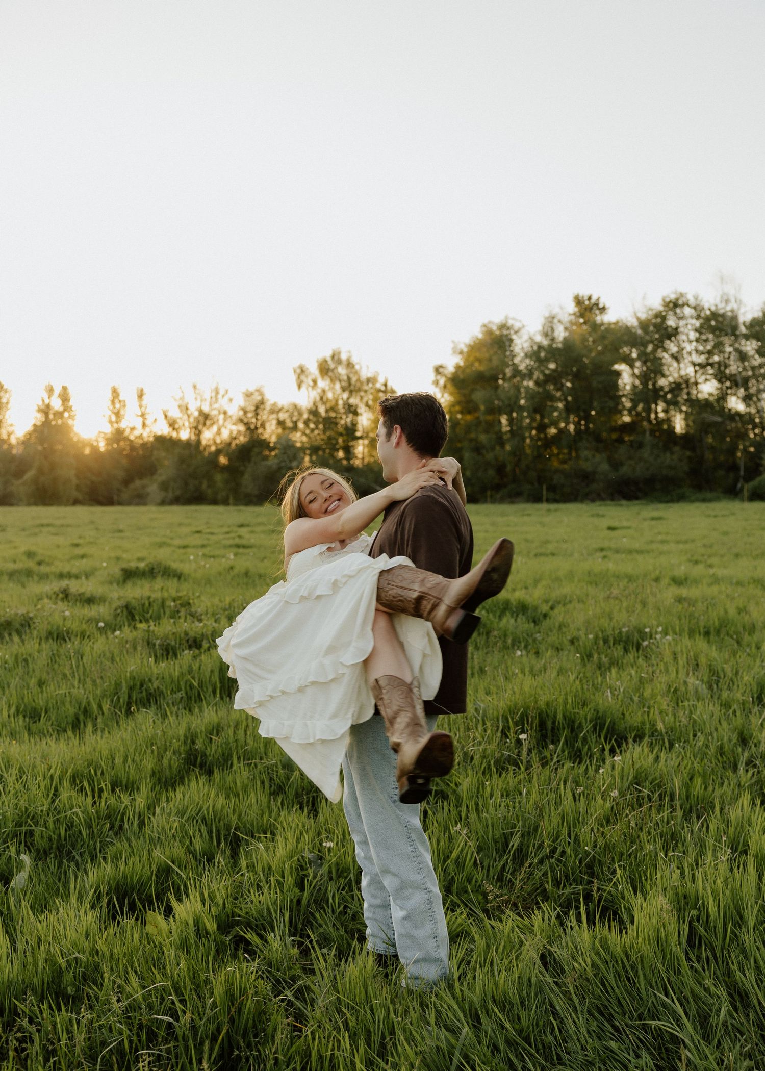 A groom lifts his bride in a white dress in a sunlit green field at golden hour.