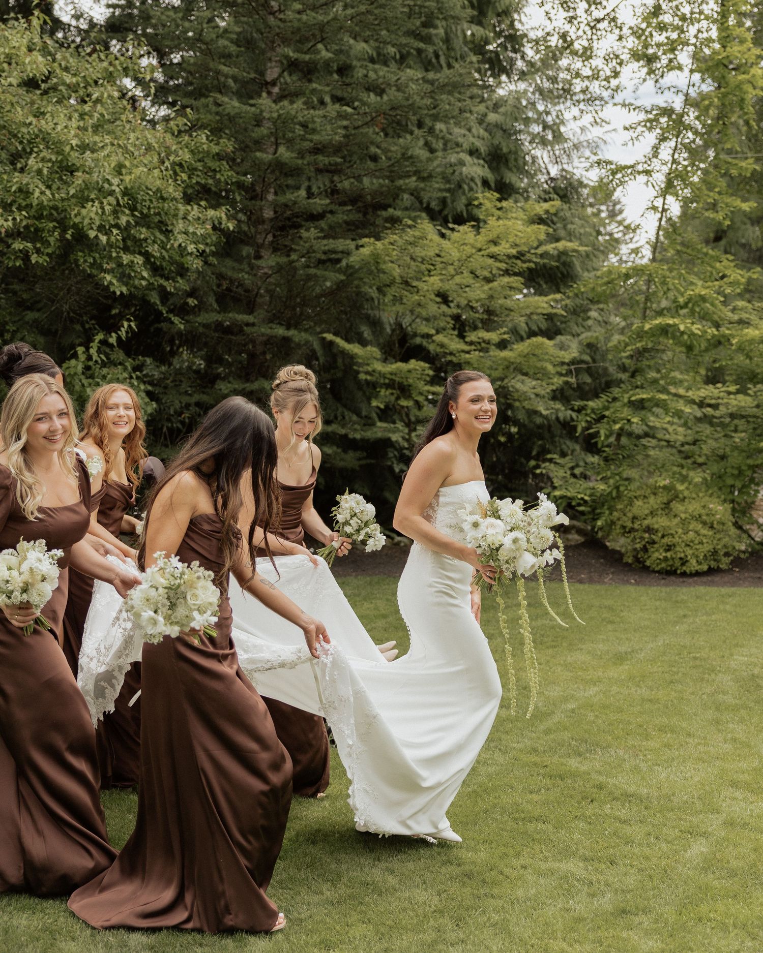 Bride in white gown walks with bridesmaids in brown satin dresses carrying white bouquets on lush green lawn.