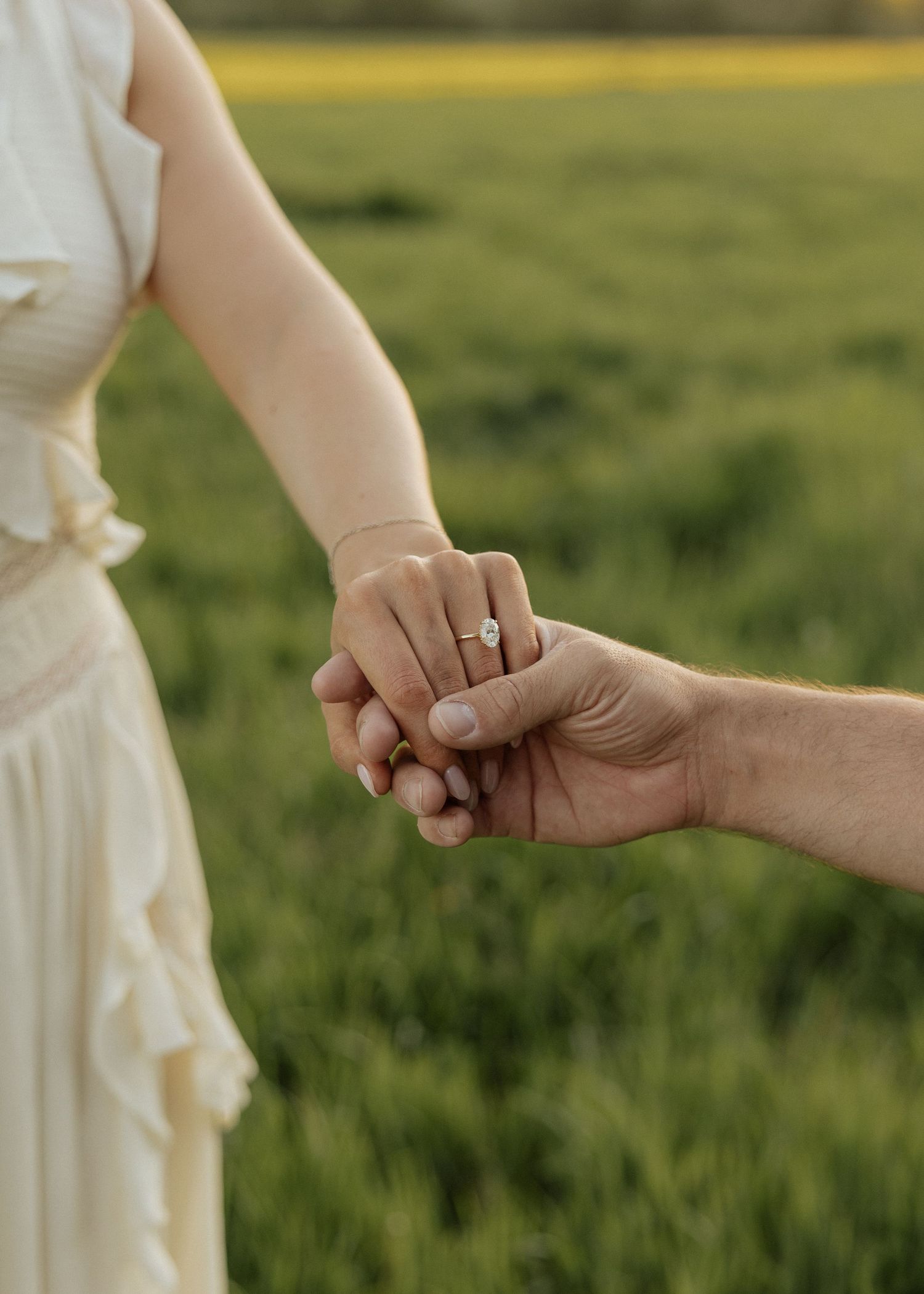 Two people holding hands in a green field, woman wearing a diamond engagement ring and white dress.