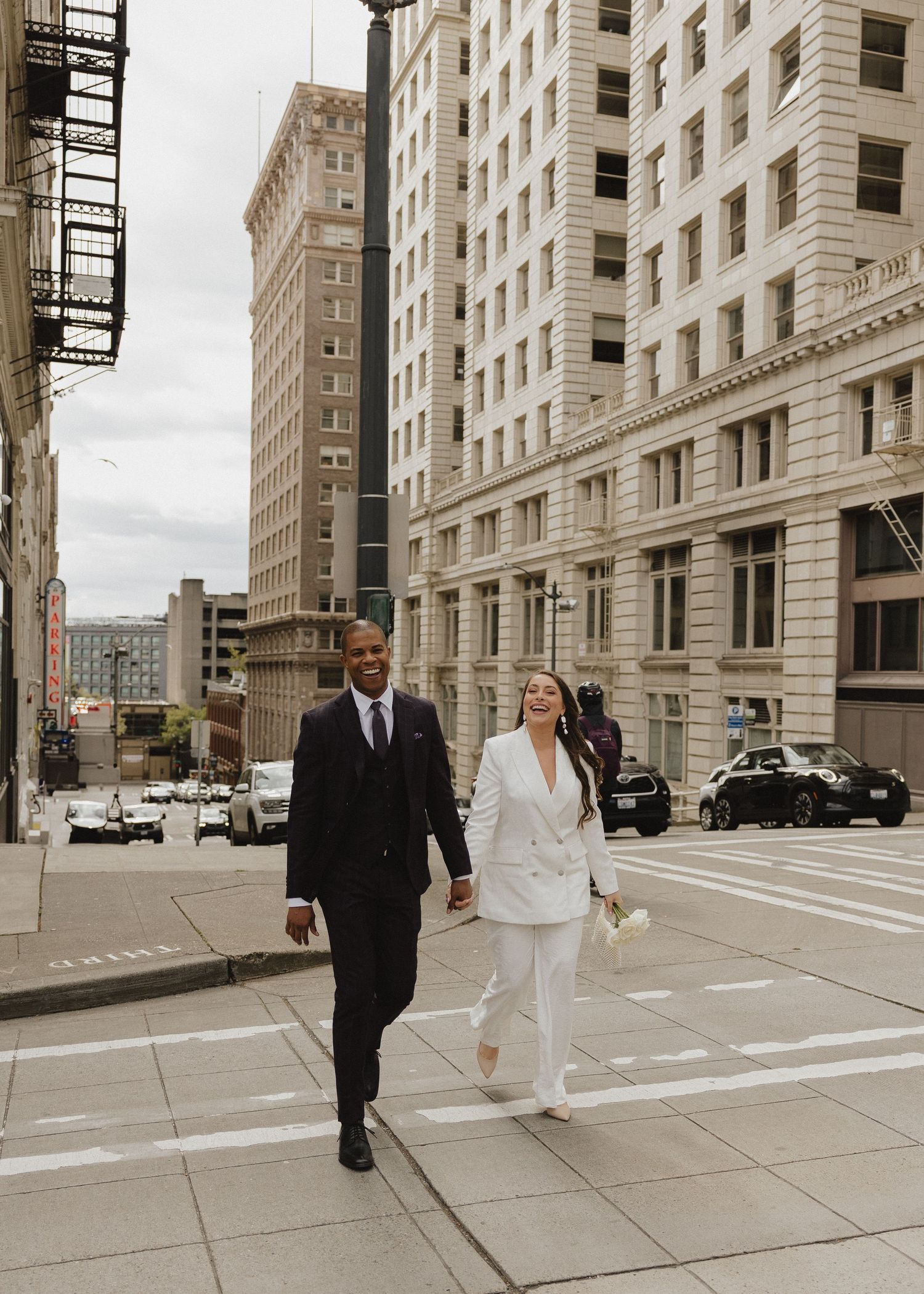 Couple in wedding attire walking hand-in-hand across a city crosswalk, groom in black suit, bride in white pantsuit.