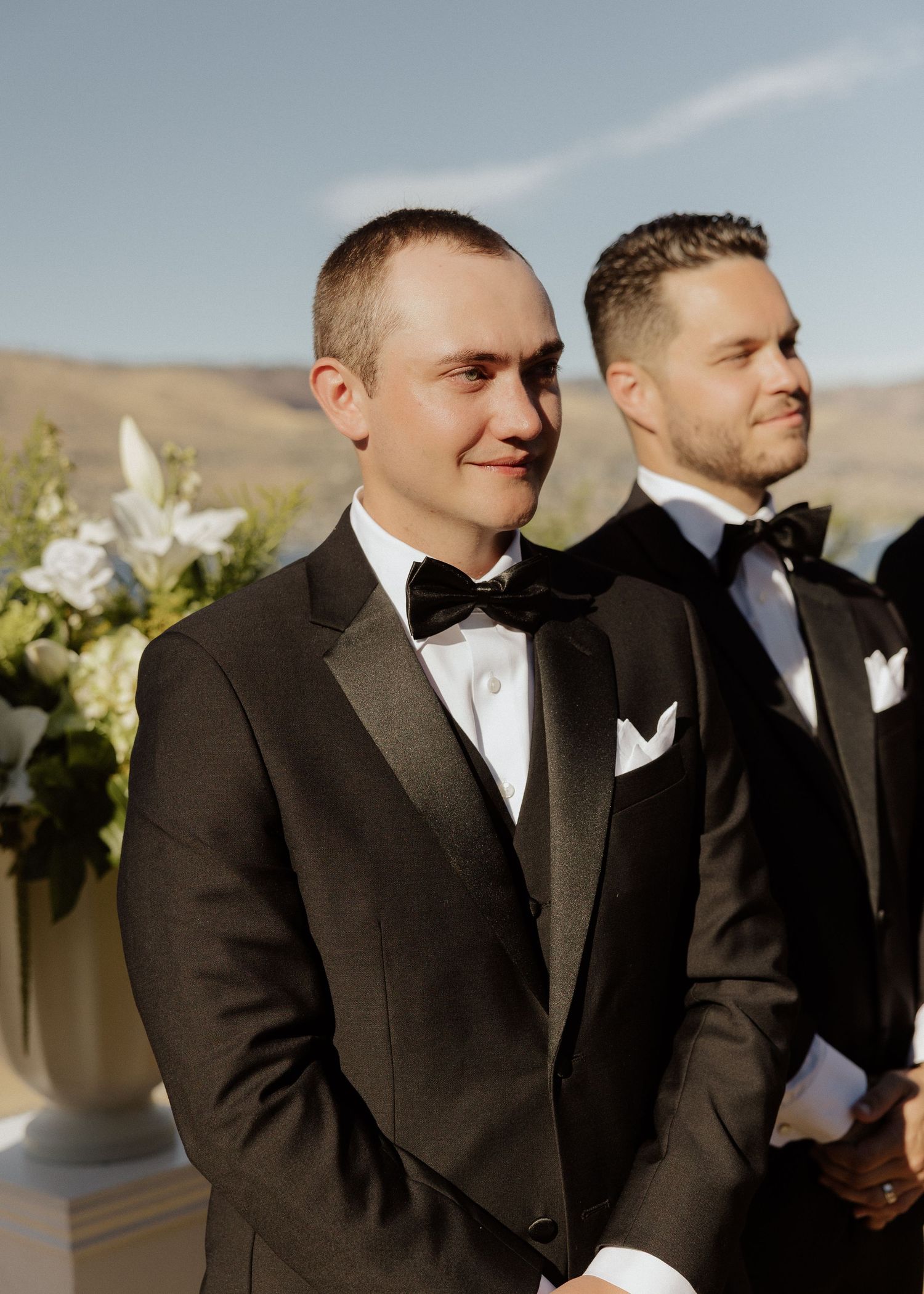 Groom in black tuxedo and bow tie stands at altar with groomsman behind him.