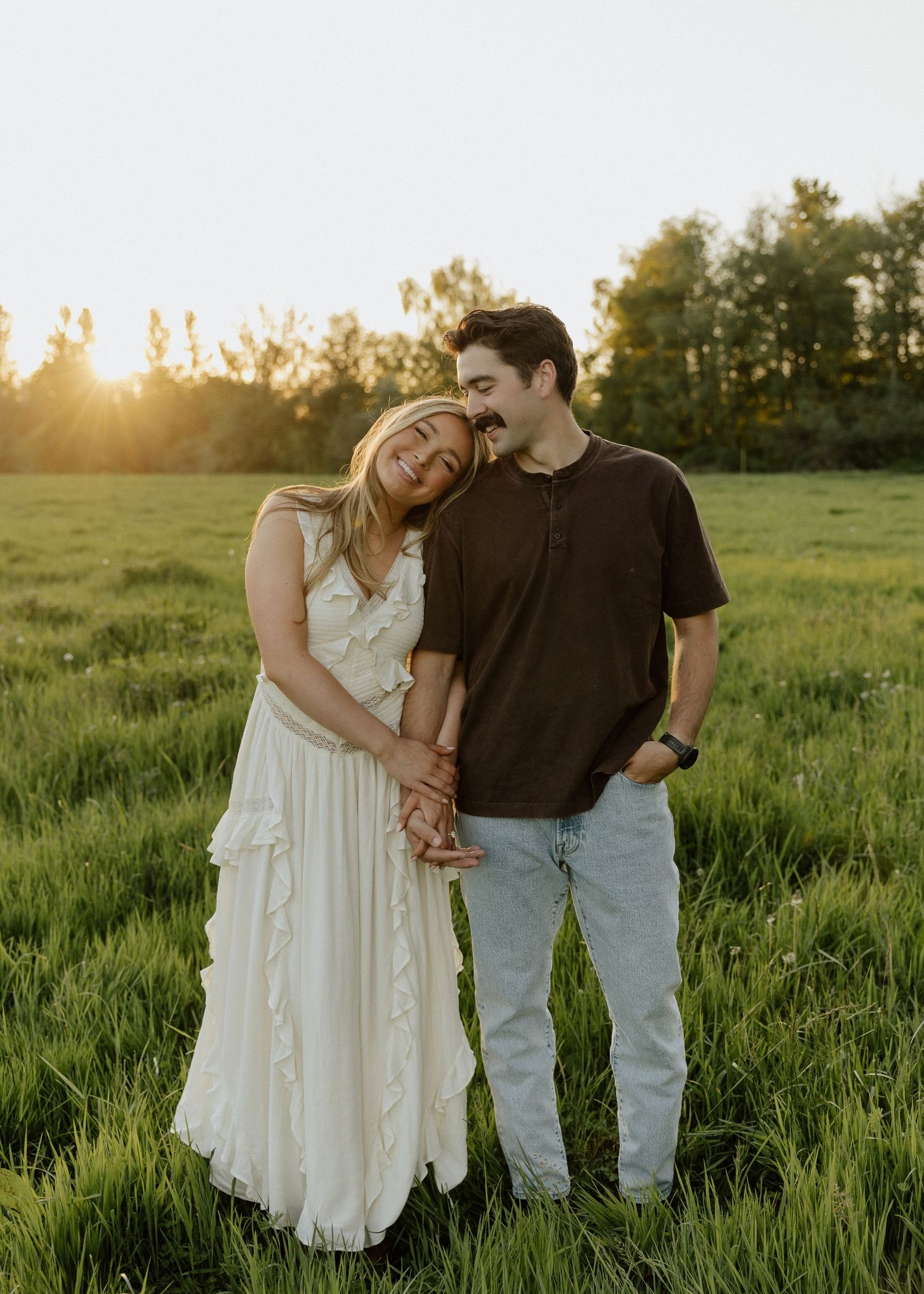 Couple laughing together in a green field at golden hour, woman in white ruffled dress, man in dark shirt and jeans.