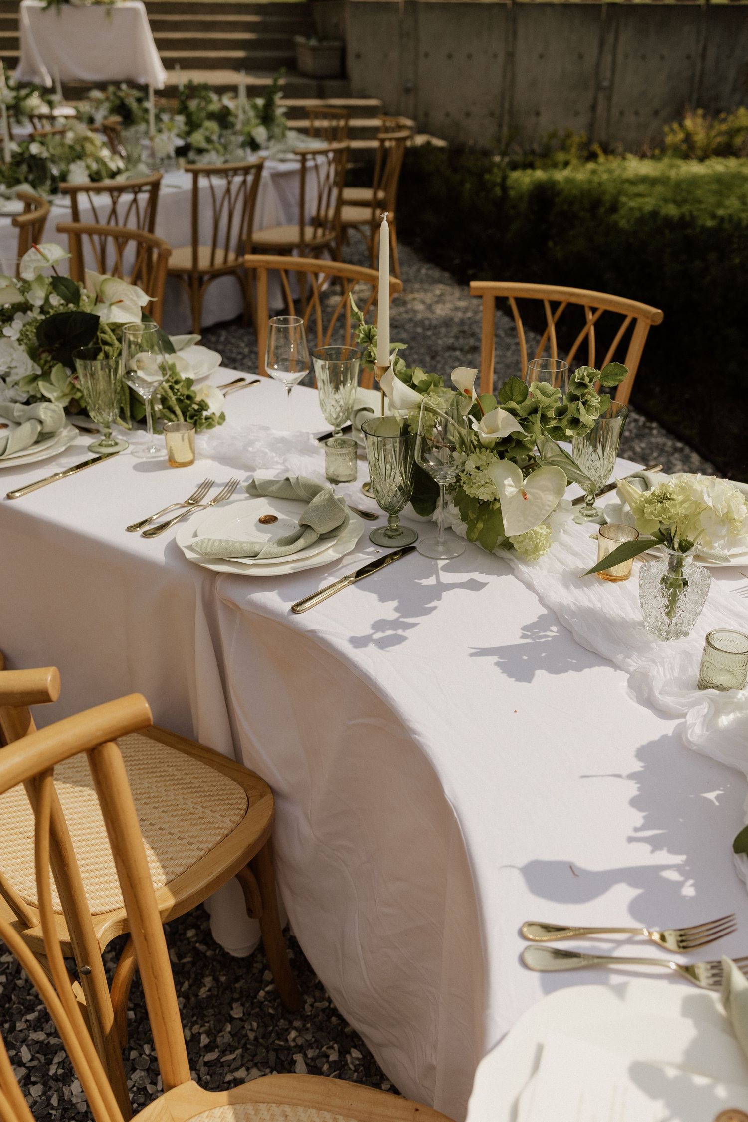 Elegant outdoor wedding reception table with white linen, greenery floral arrangements, gold cutlery, and wooden chairs.
