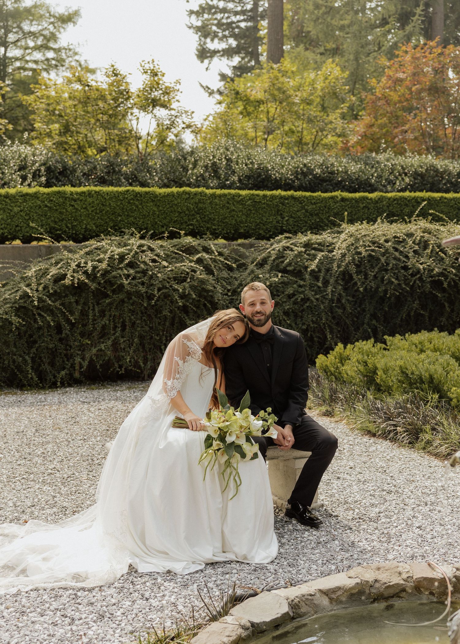 Bride and groom pose in a lush garden, bride leaning on groom's shoulder holding a white floral bouquet.
