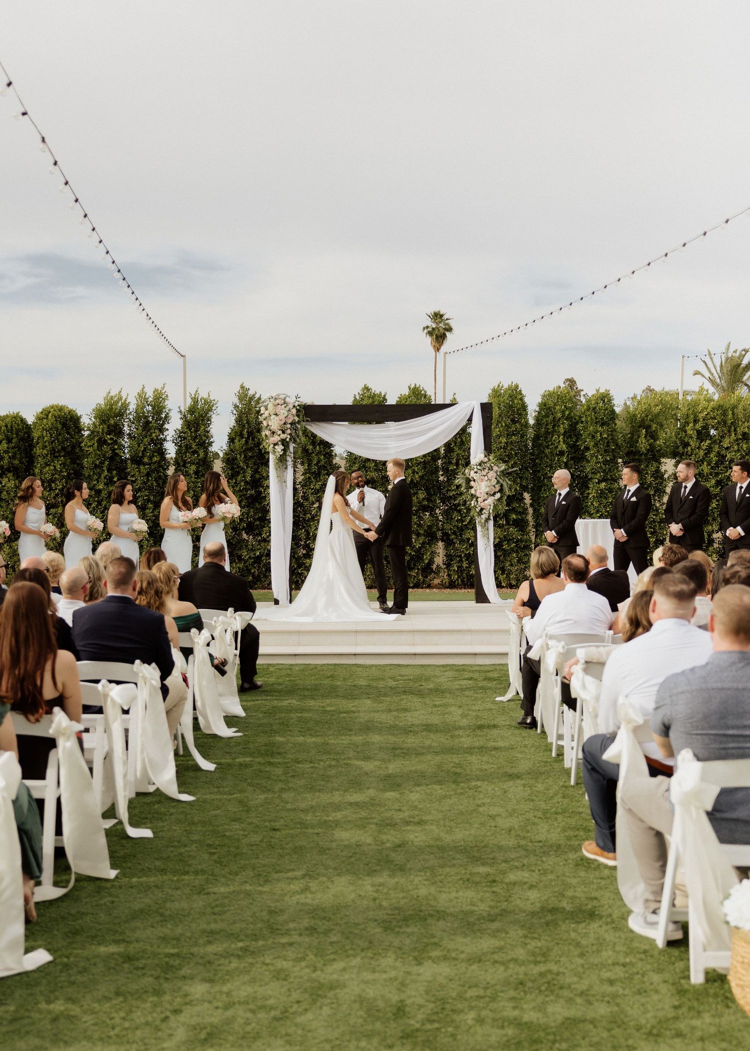 Elegant outdoor wedding ceremony on green lawn with white arch draped in fabric and floral arrangements.