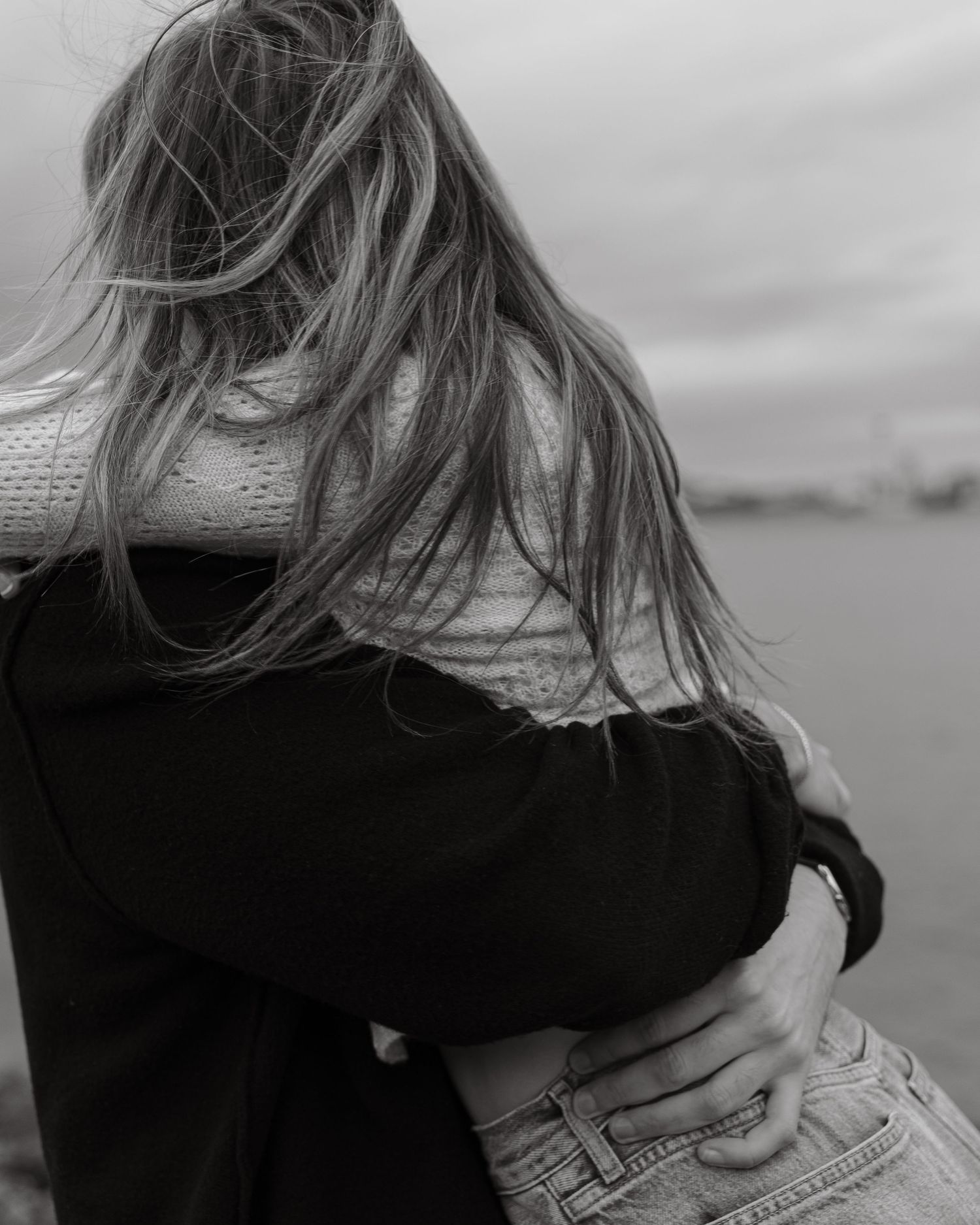 A close-up of someone embracing another person from behind with windswept hair against an overcast sky.