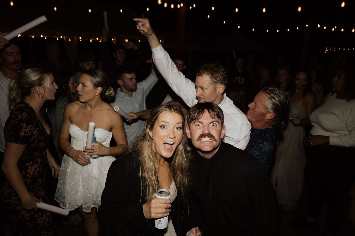 Wedding party guests celebrate on dance floor with sparklers under string lights at outdoor reception.