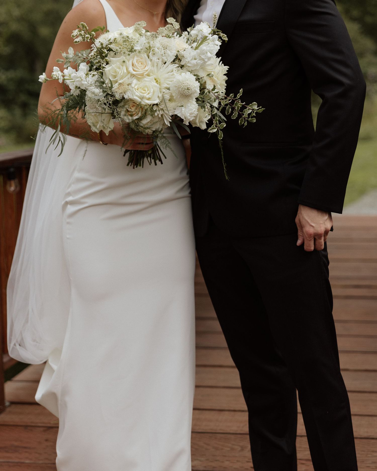White wedding bouquet detail with bride and groom on rustic wooden deck.