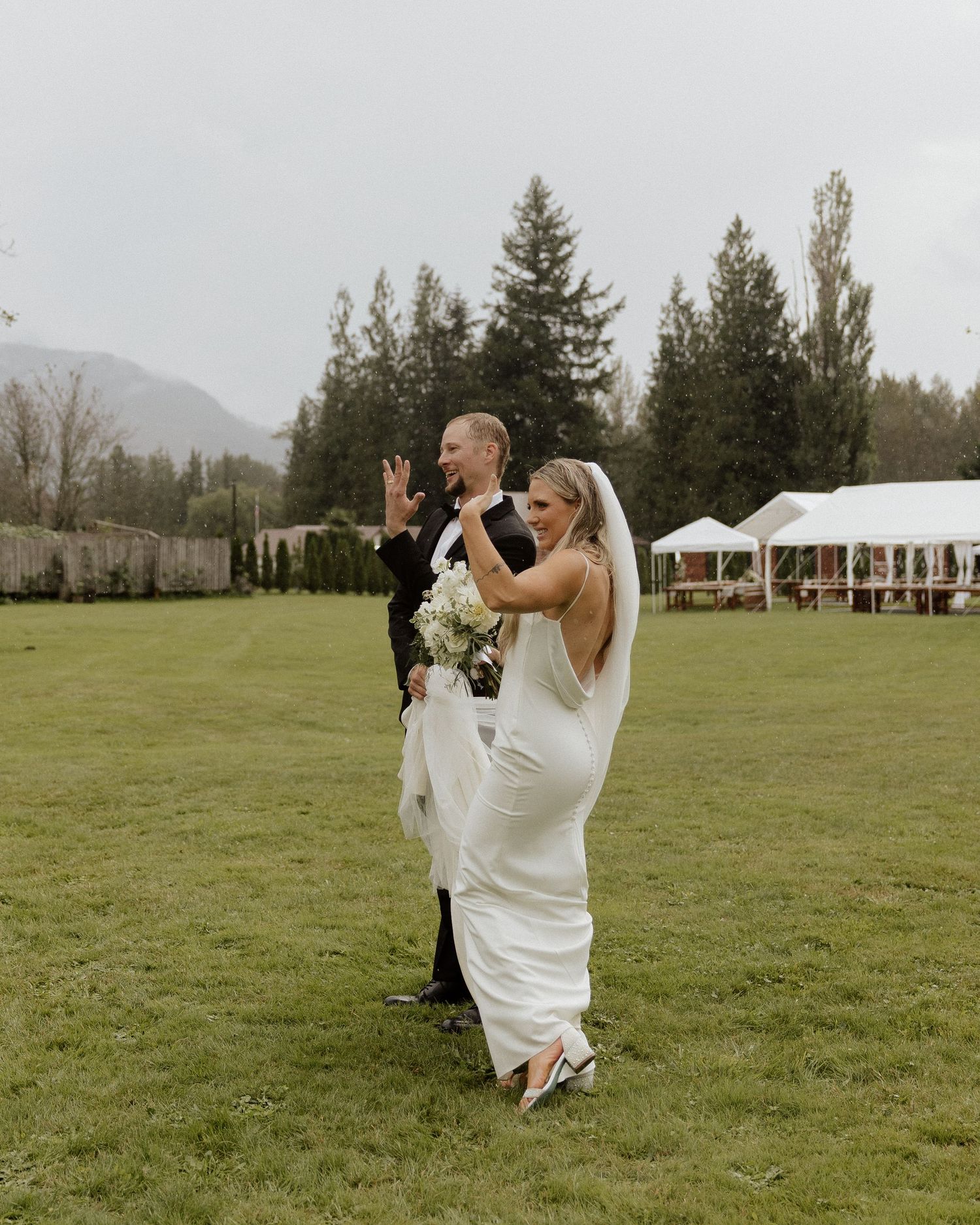 Newlyweds celebrate with energetic poses on grassy field with mountain backdrop.
