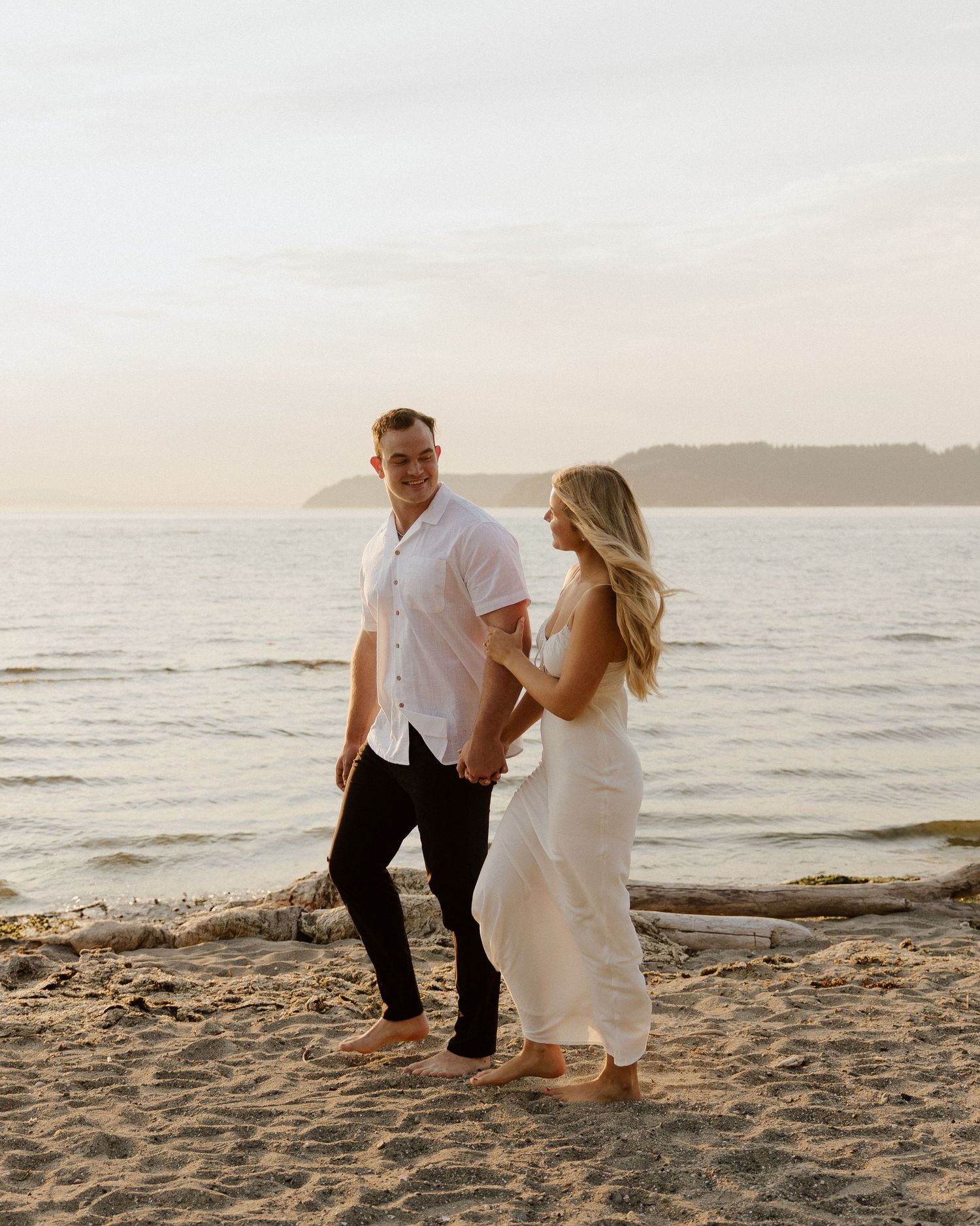 A couple walks along a beach at sunset wearing white outfits against a serene ocean backdrop.