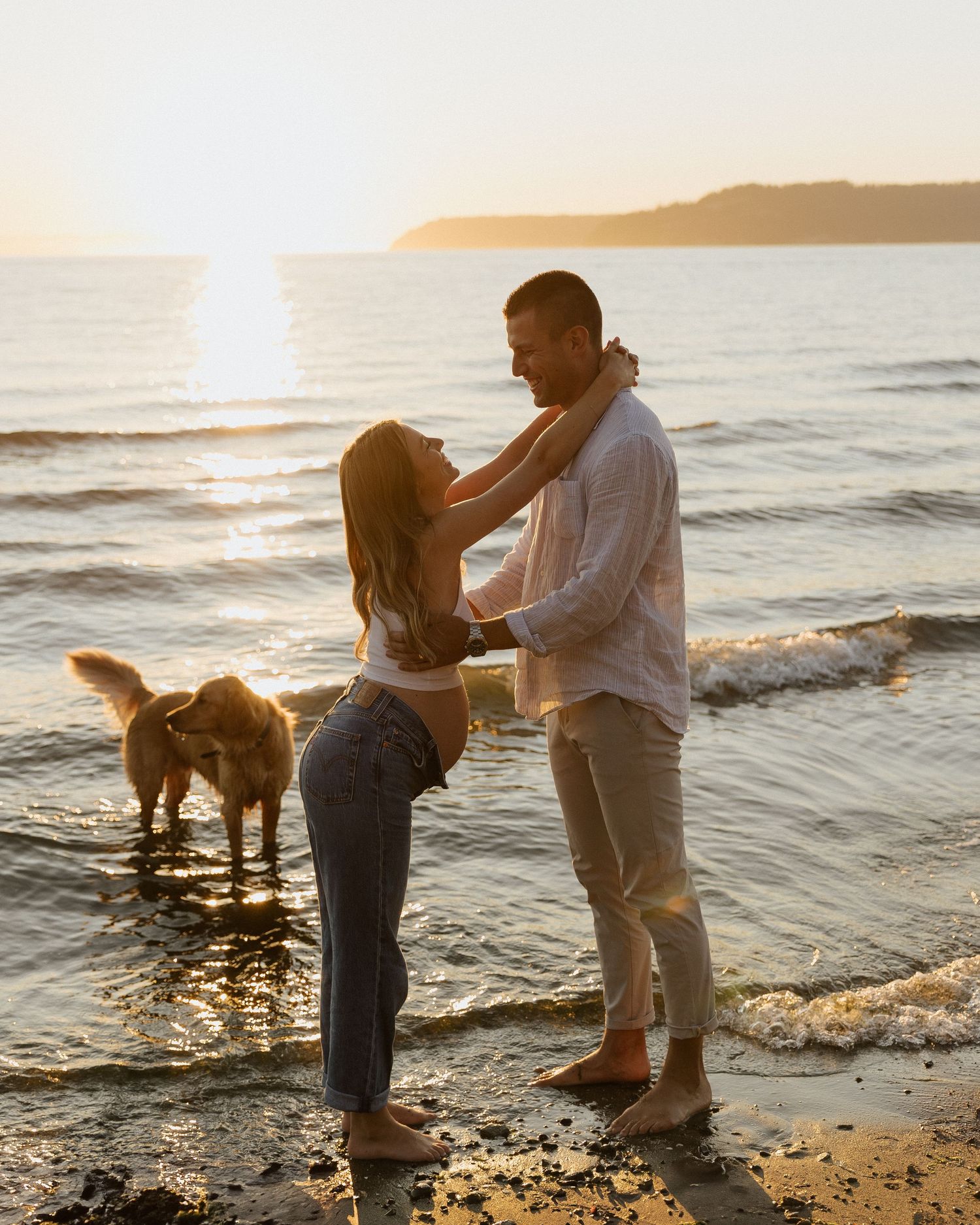 Couple embraces on beach at sunset with golden retriever beside them.