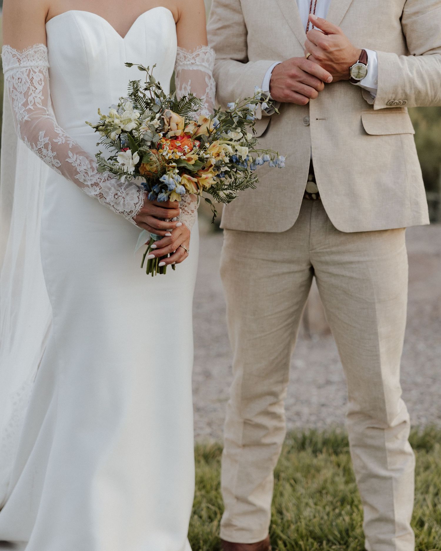 Detail shot of bride holding bouquet next to groom in beige suit against natural backdrop.