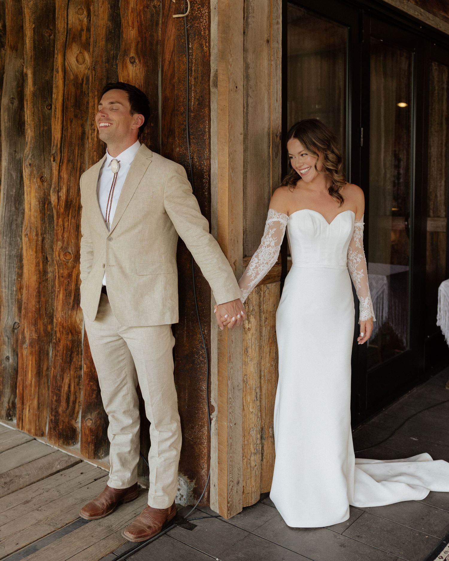 Bride in white gown and groom in beige suit pose separated by rustic wooden wall for first look photo.