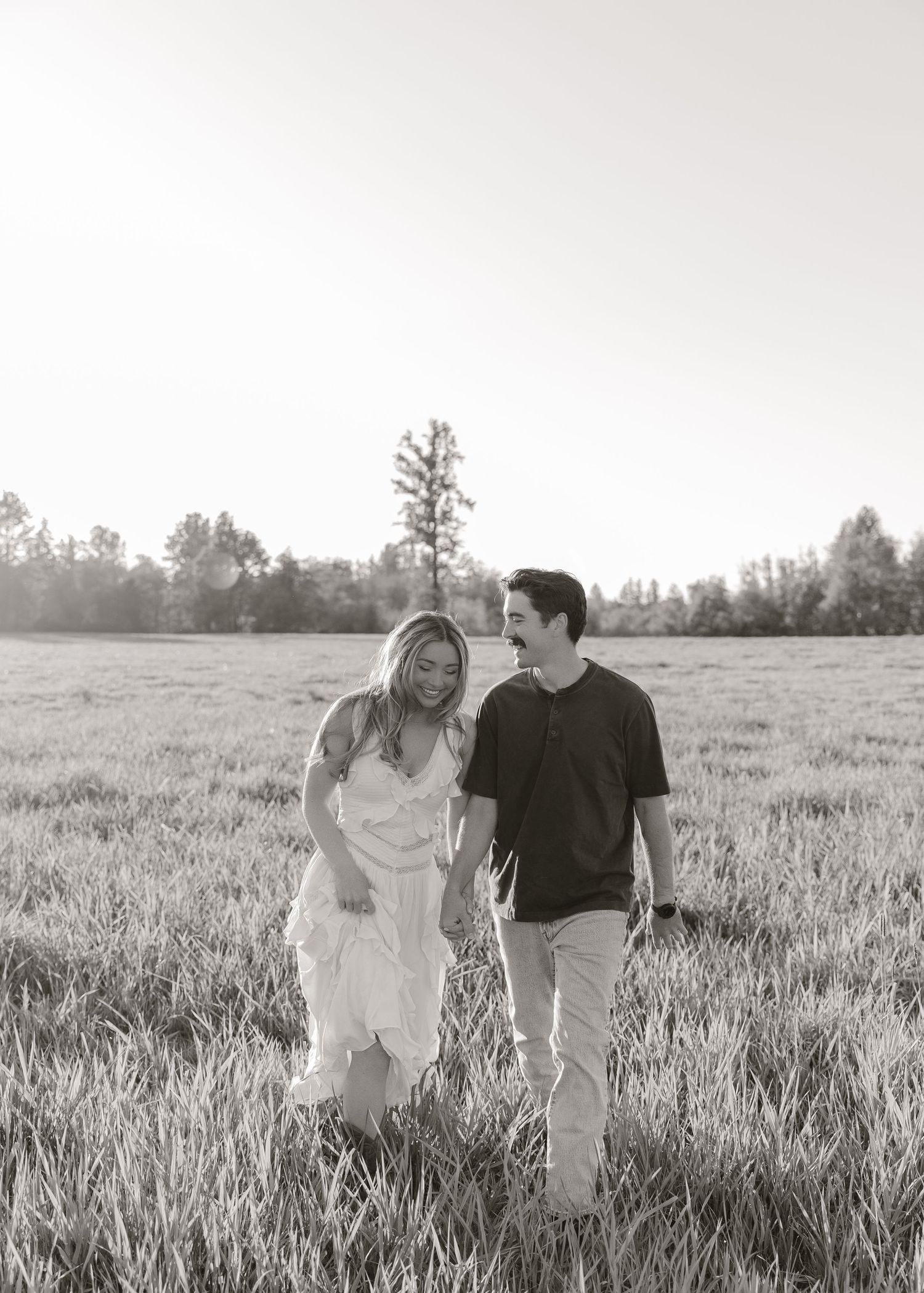Black and white photo of a smiling couple walking hand in hand through a sunlit grassy field with trees in the background.