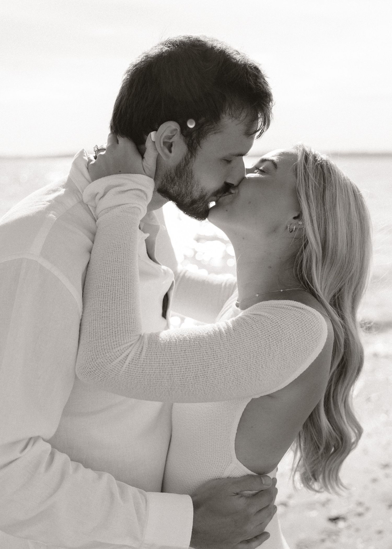 Couple sharing a romantic kiss on the beach in black and white, both dressed in white.