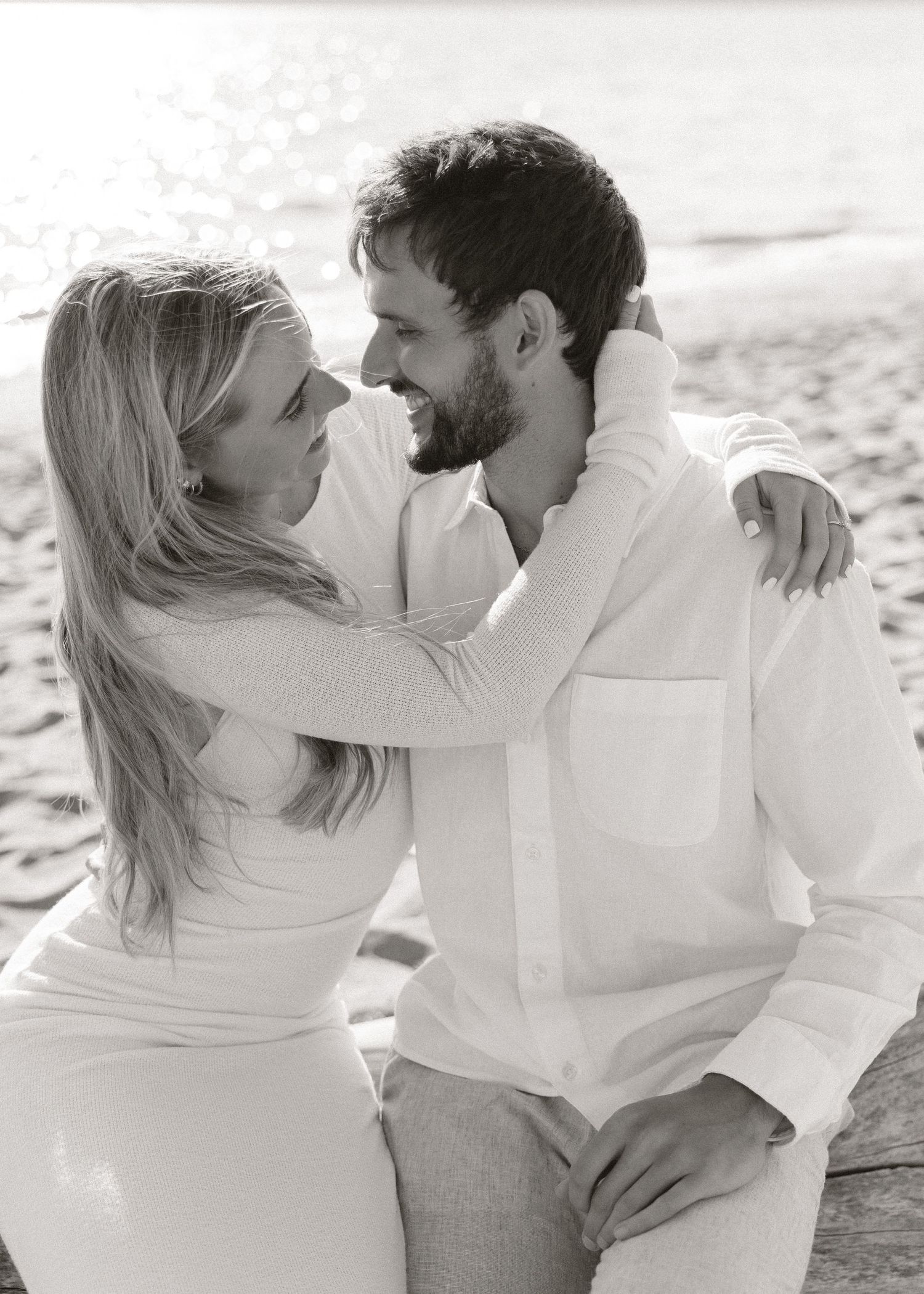 Black and white photo of a smiling couple in white outfits embracing on a beach, gazing lovingly at each other.