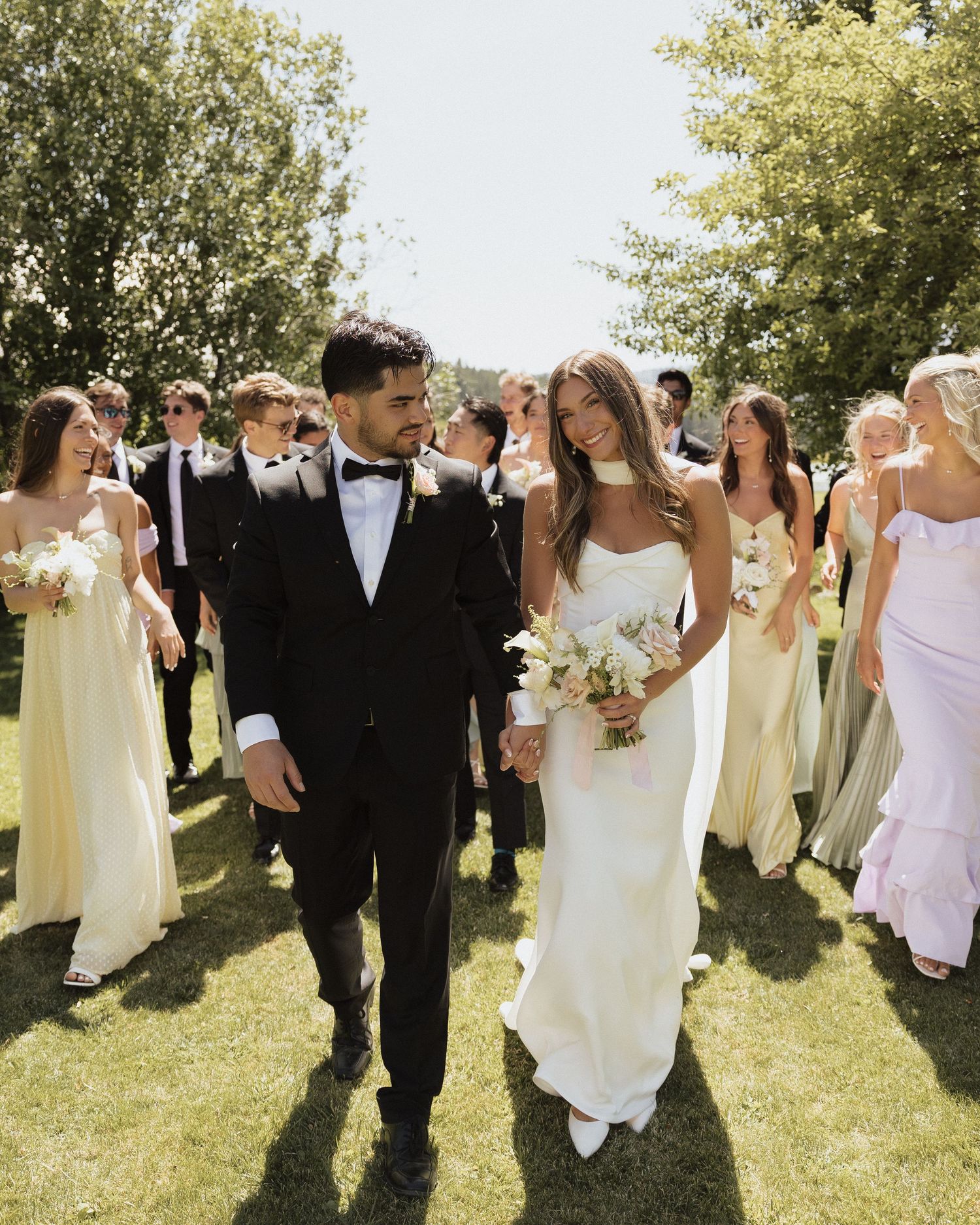 Bride and groom walk down the aisle surrounded by wedding party in yellow dresses and black suits outdoors.