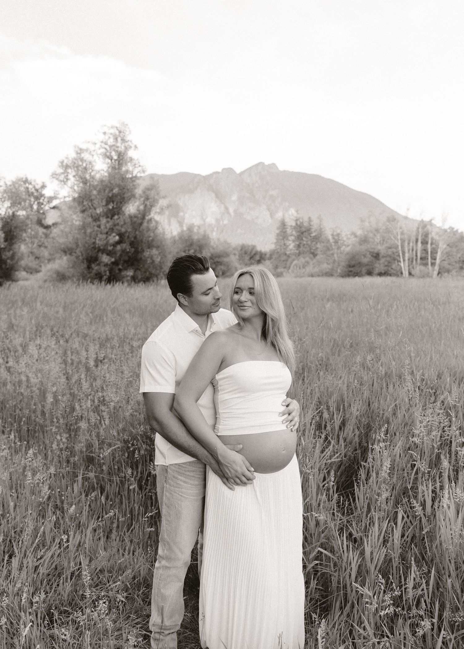 Black and white maternity photo of a couple embracing in a field with mountains in the background.
