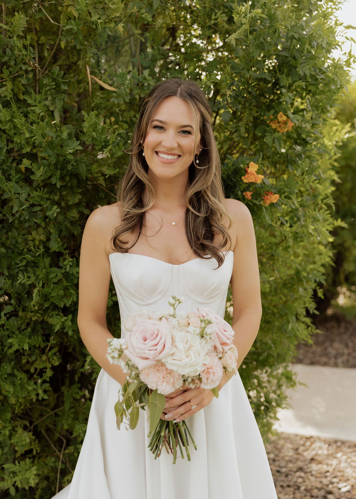 Smiling bride in strapless white gown holding pink rose bouquet, posing outdoors among lush green hedges.