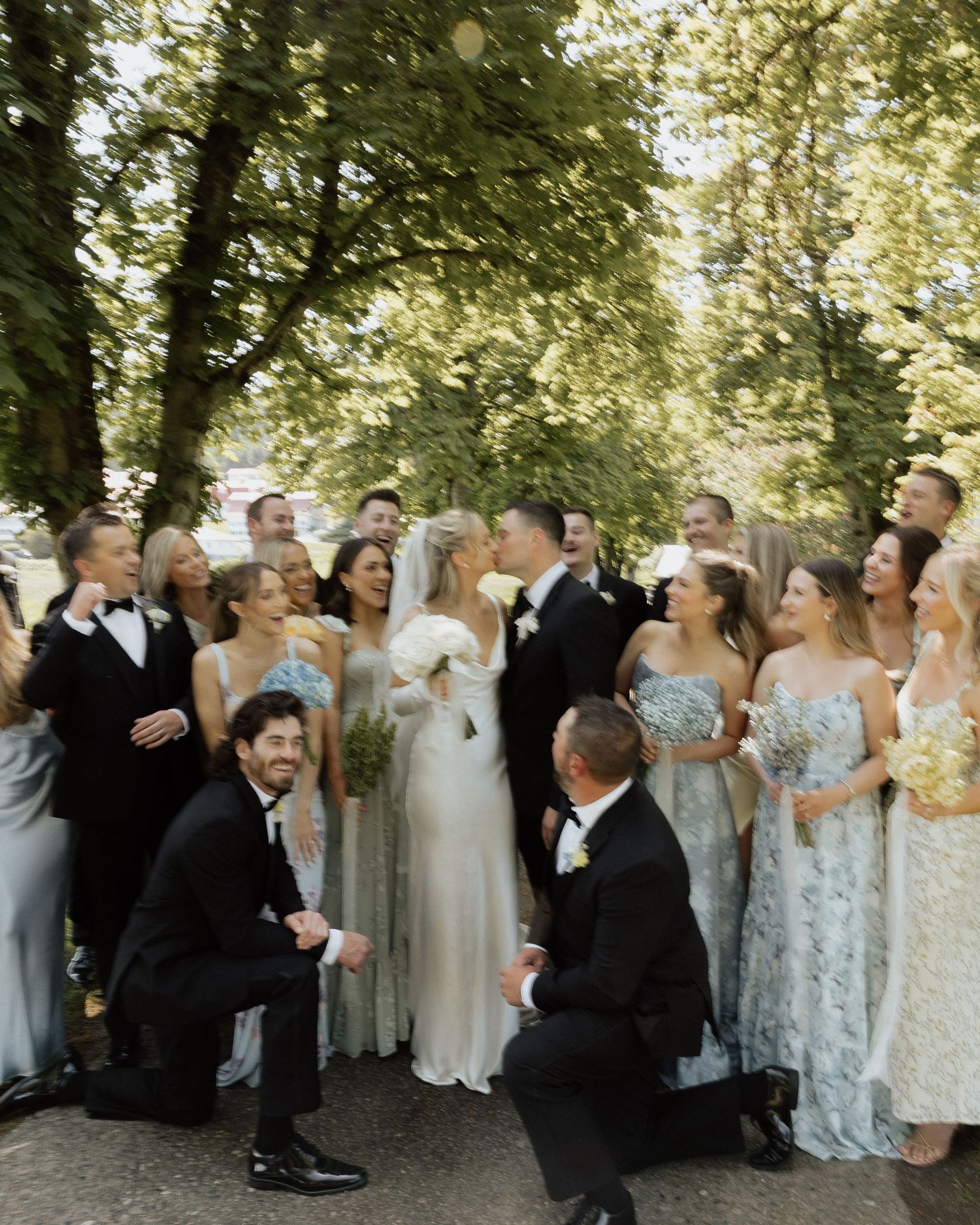 Large wedding party poses outdoors under green trees, bride and groom share a kiss surrounded by bridesmaids and groomsmen.