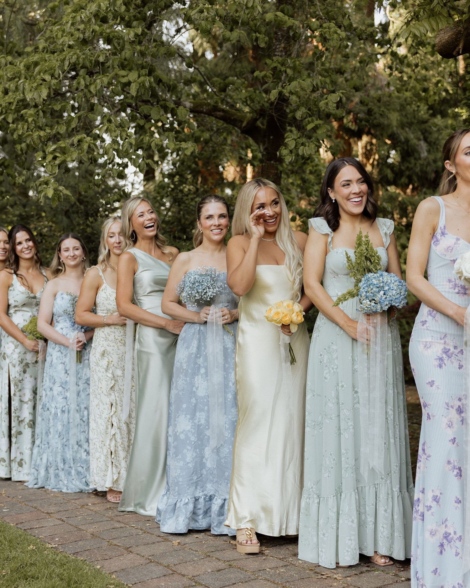 Bridesmaids in mismatched floral and pastel maxi dresses holding blue hydrangea bouquets laughing outdoors at a garden wedding.