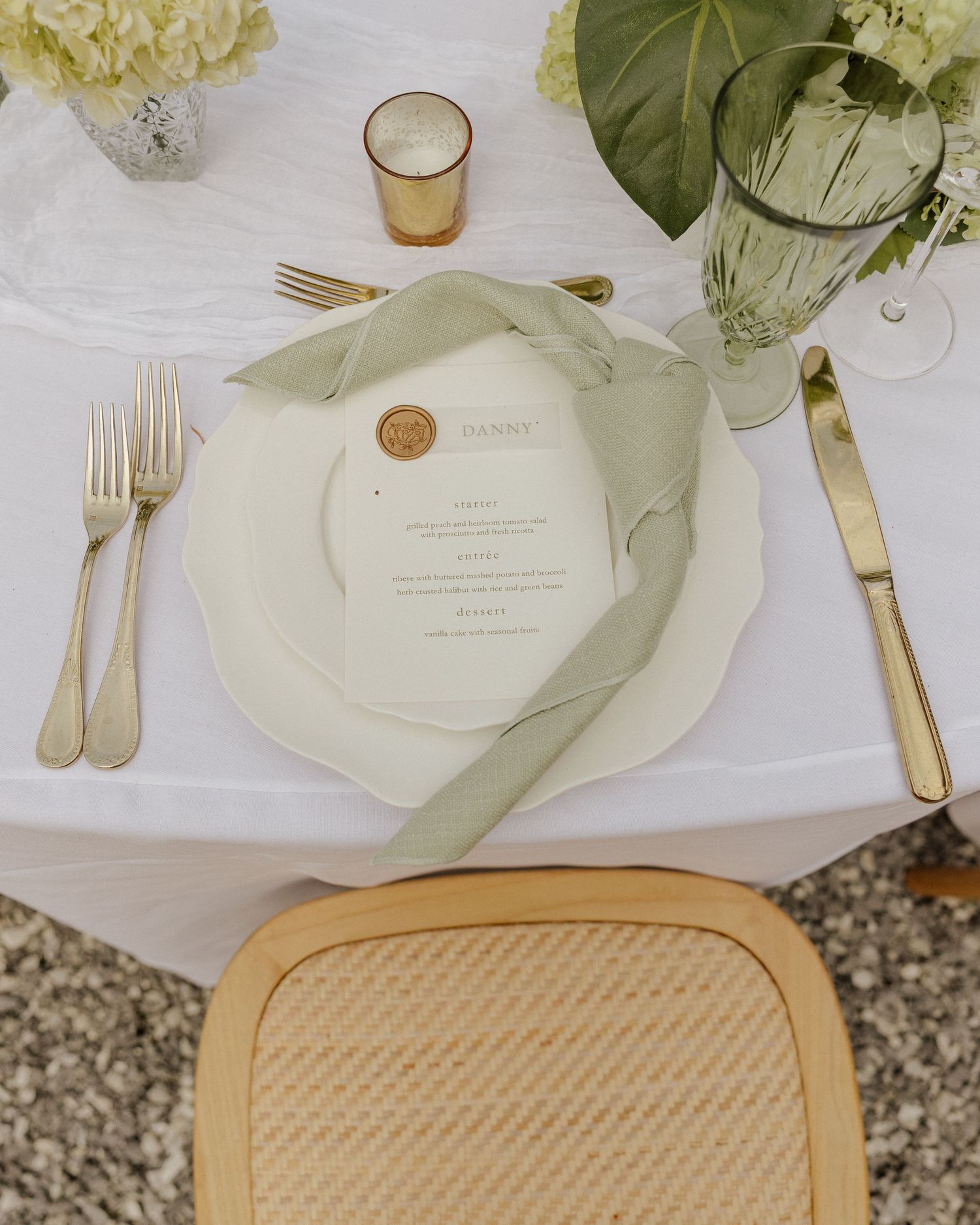 Elegant wedding place setting with gold cutlery, sage green ribbon, menu card, and floral centerpiece on white linen.