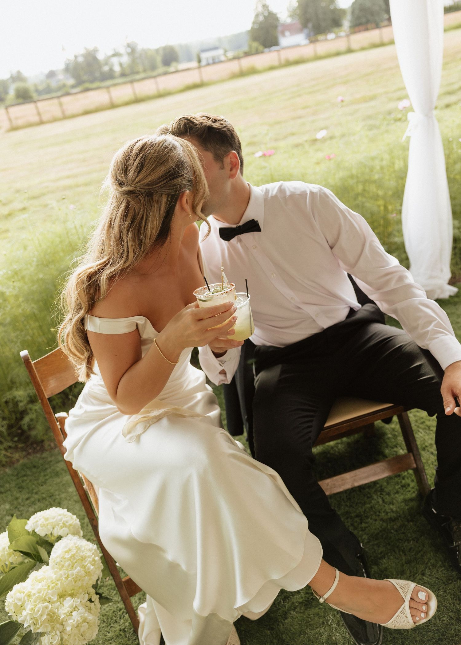 Bride and groom sit together outdoors sharing a drink, groom kissing bride on the head during wedding reception.