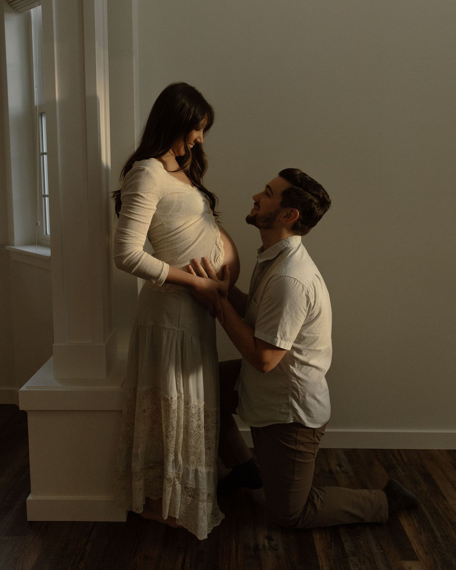 Romantic maternity photo with couple in white against neutral wall and hardwood floors.