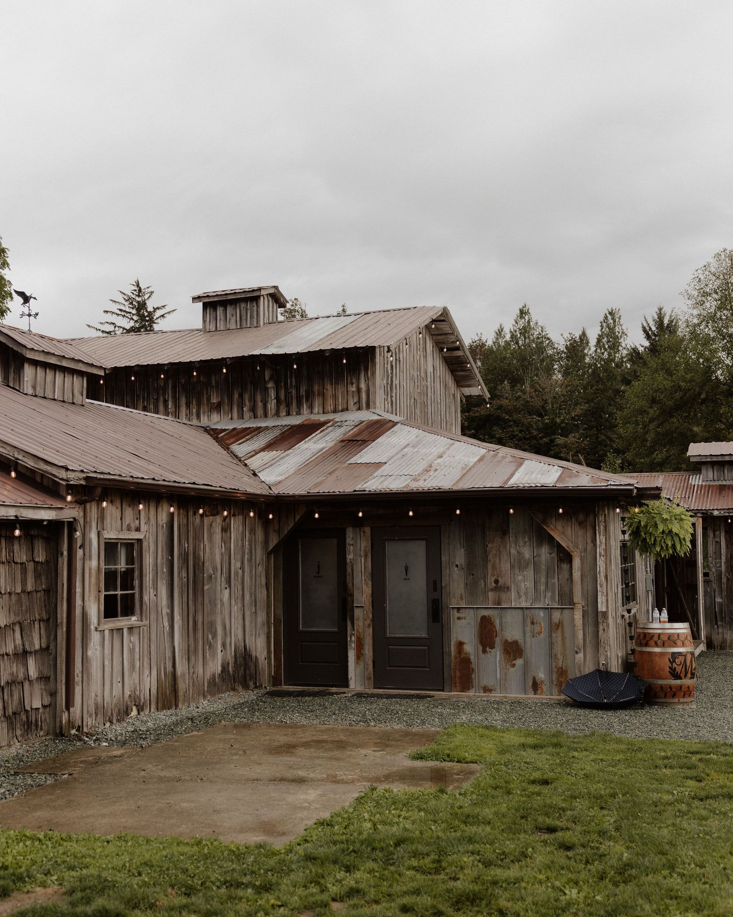 A rustic wooden barn with string lights and weathered siding sits against a forested backdrop.