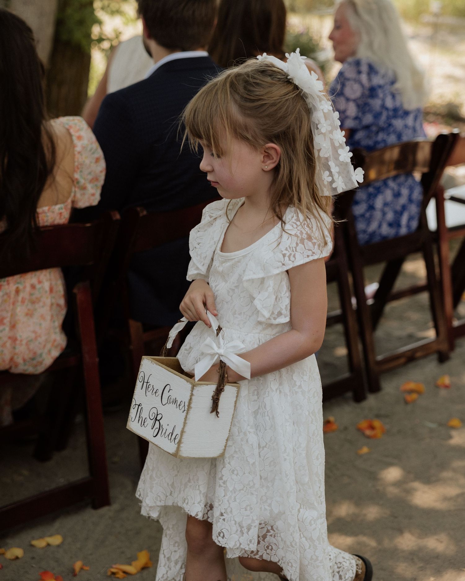Young flower girl in white lace dress holds basket at outdoor wedding ceremony.