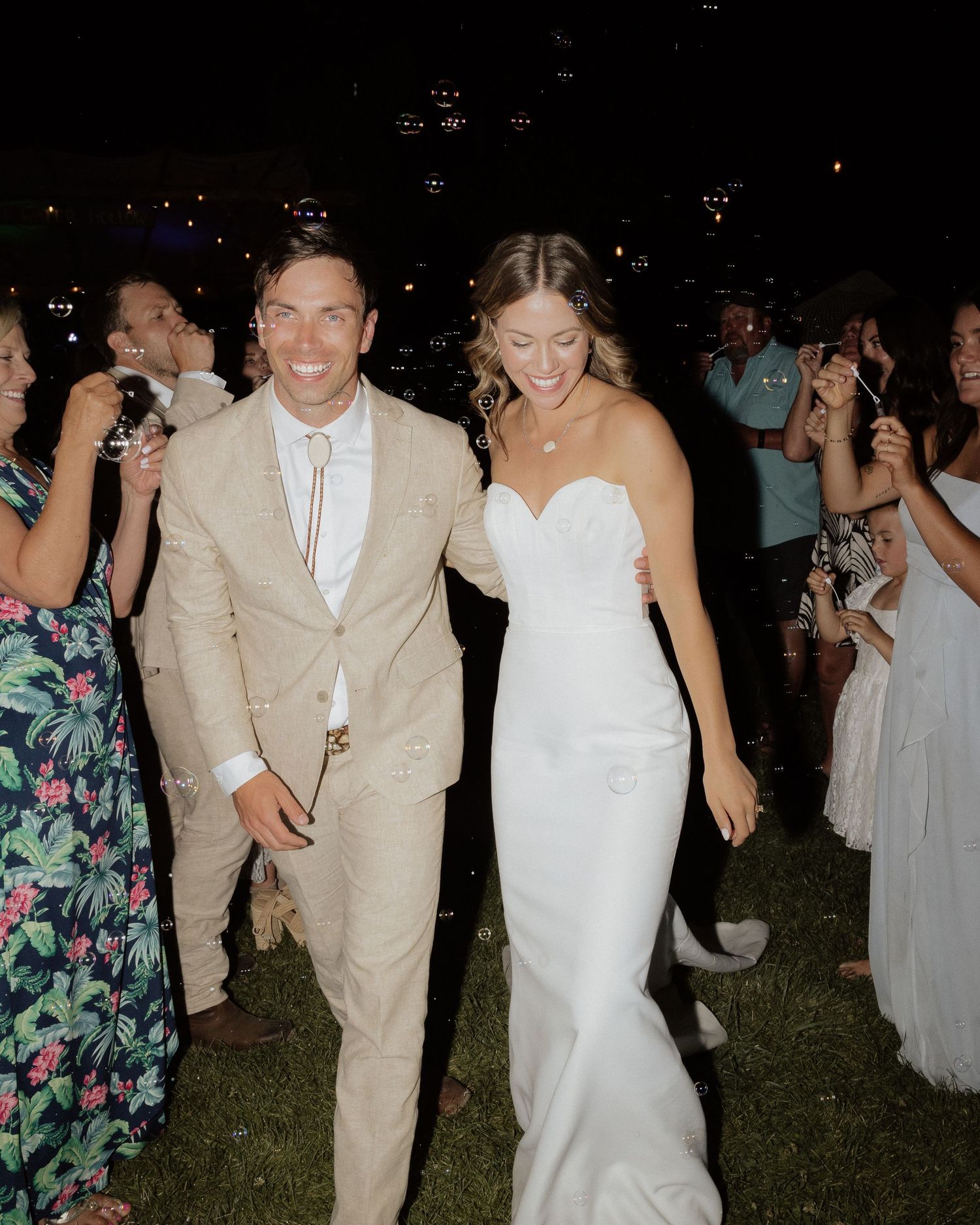Newlyweds walking together at night celebration with guests cheering in background.
