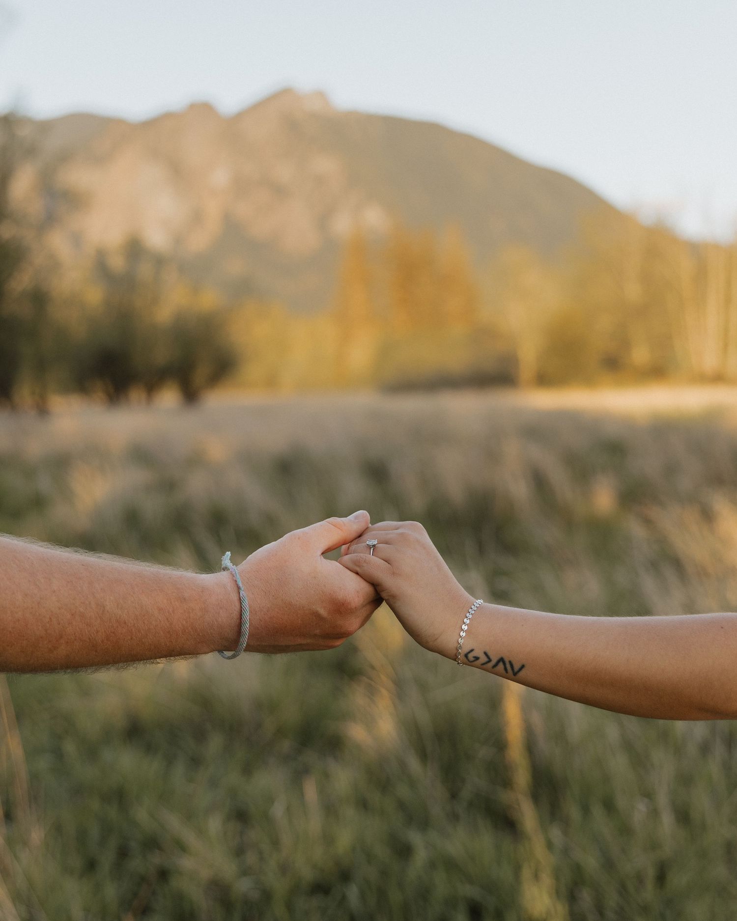 Hands clasped together with tattoo visible against mountain landscape at sunset.