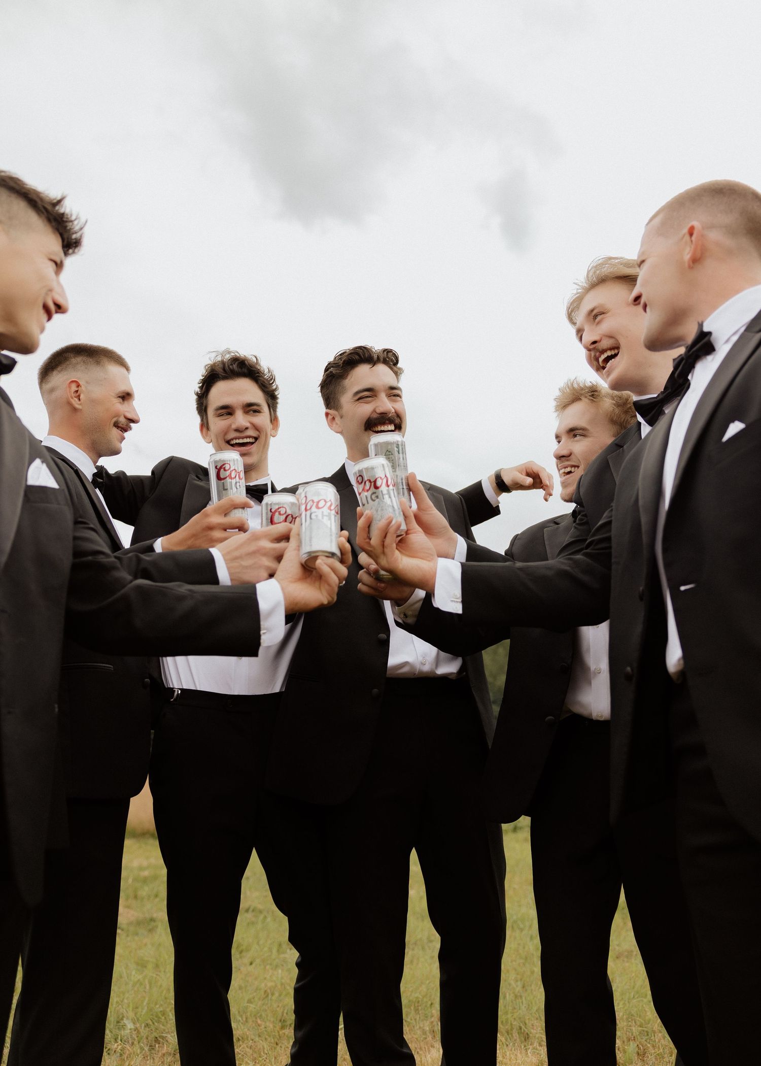 Groomsmen in black tuxedos toasting with cans outdoors on a cloudy day at a wedding celebration.