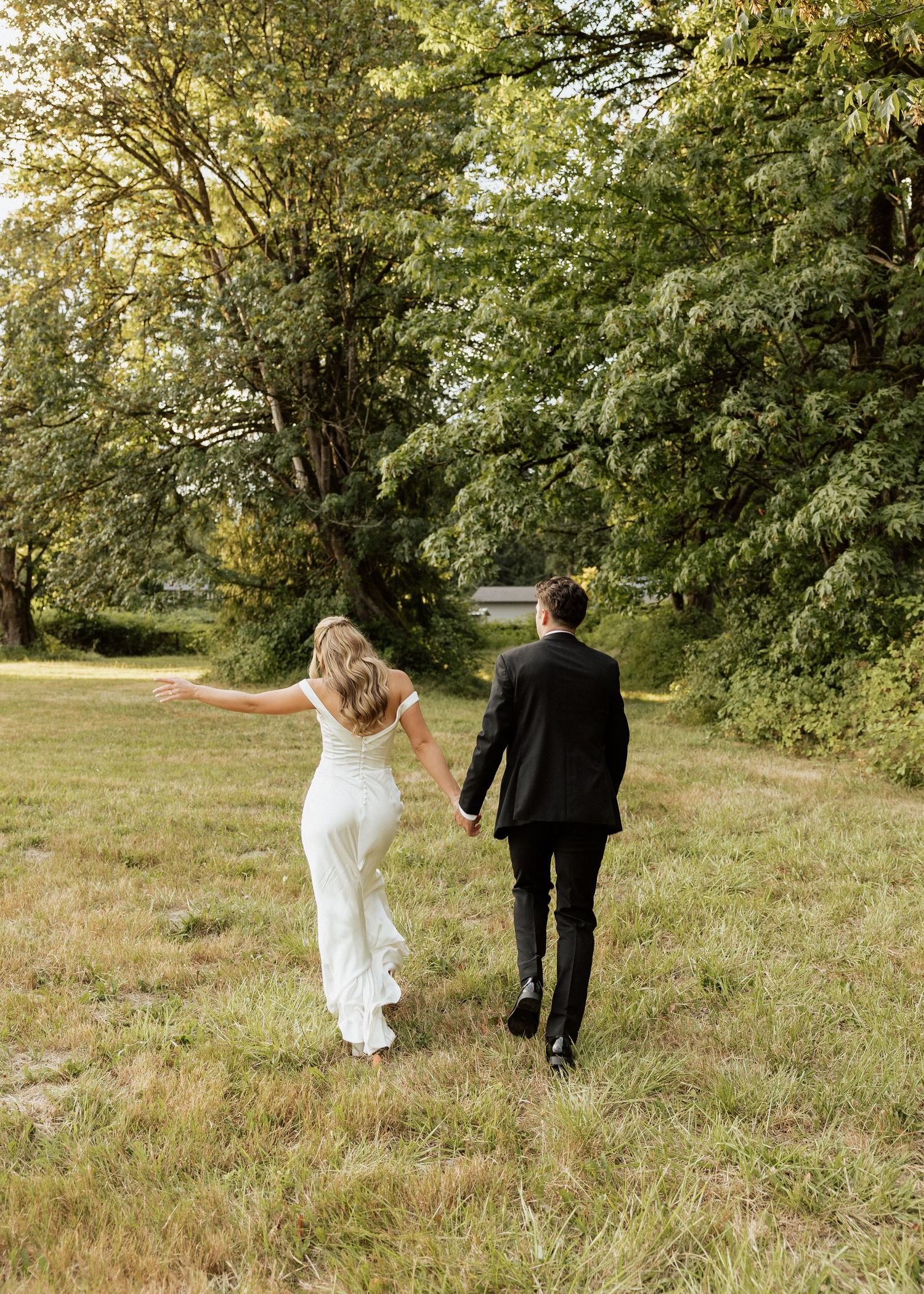 Bride and groom walk hand-in-hand through a lush green meadow surrounded by tall trees on their wedding day.
