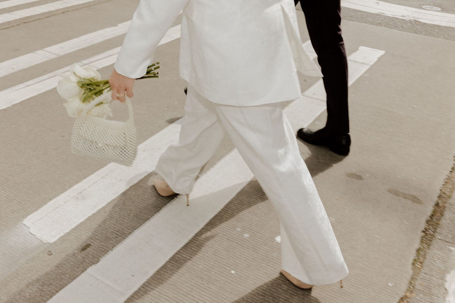 Person in white suit carrying flowers crosses a zebra crossing, elegant bridal street style photo.