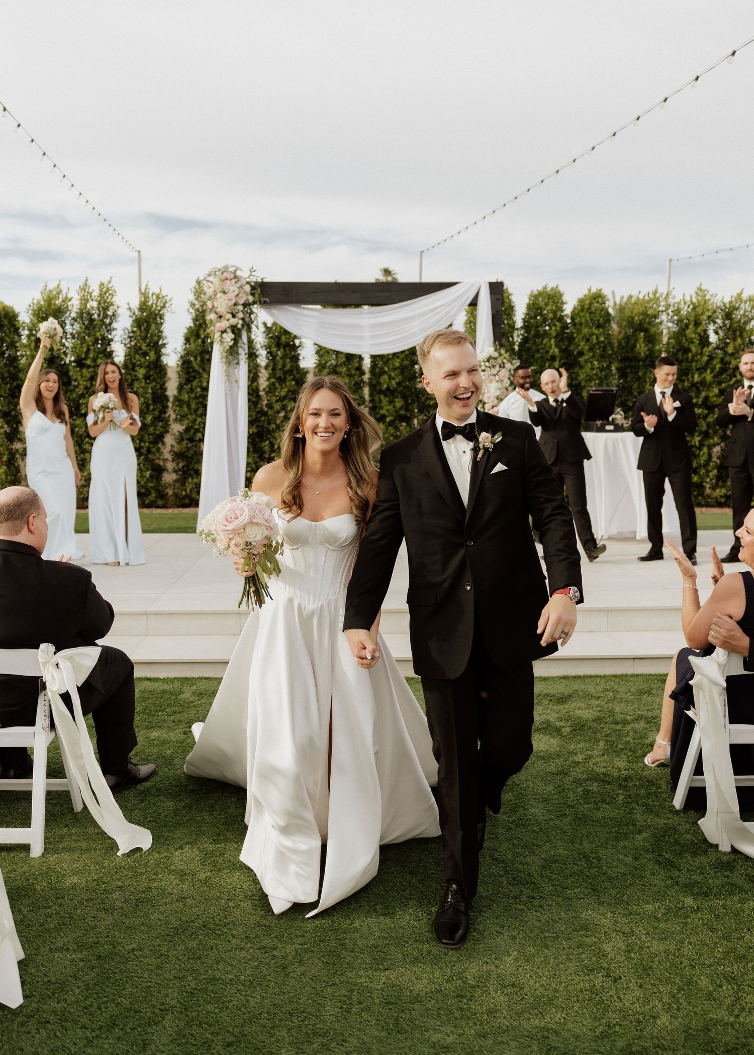 Happy newlyweds walk down aisle after outdoor wedding ceremony on green lawn with string lights.