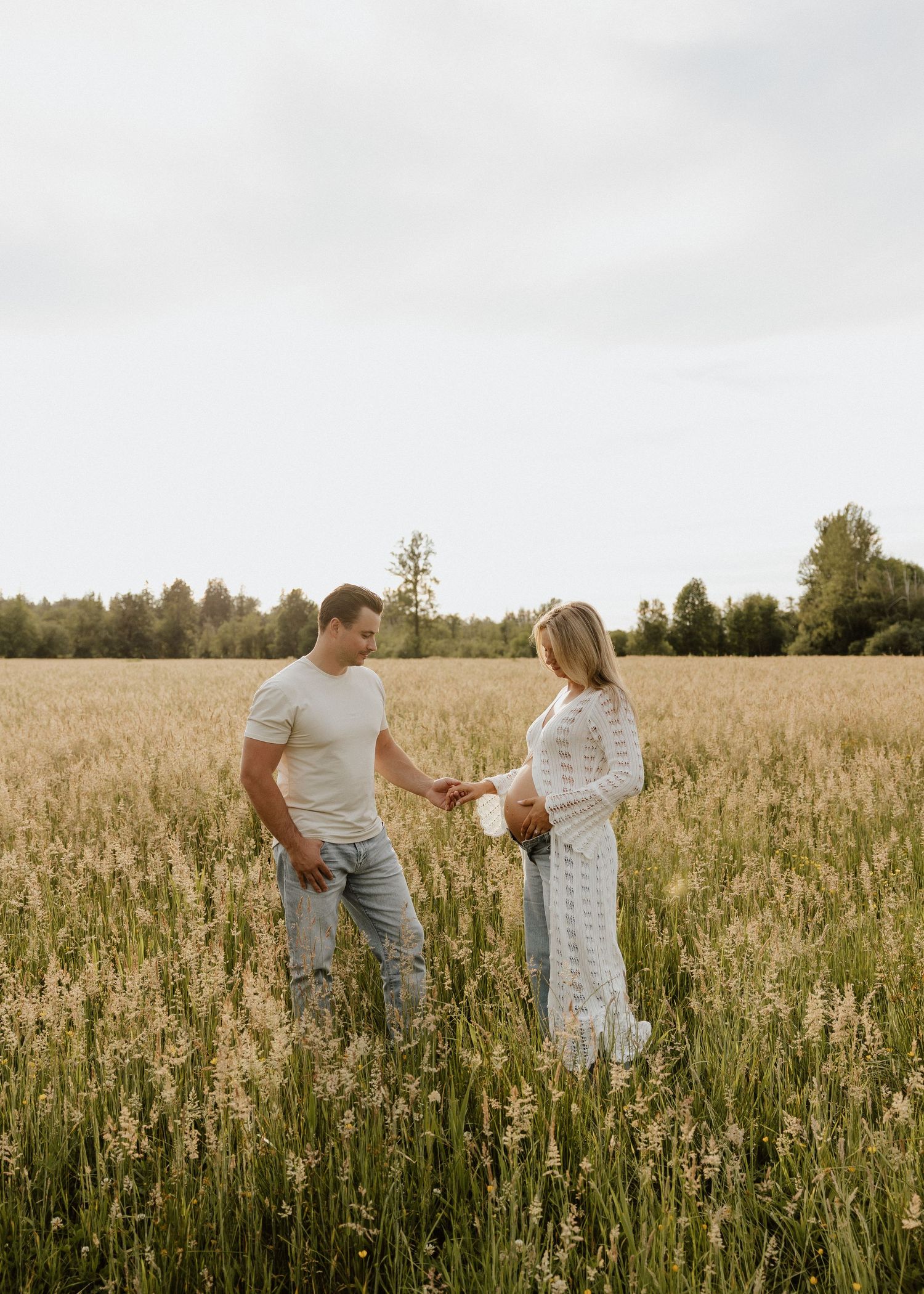 A couple stands in a golden field, the man handing a flower to a pregnant woman in a white lace dress.