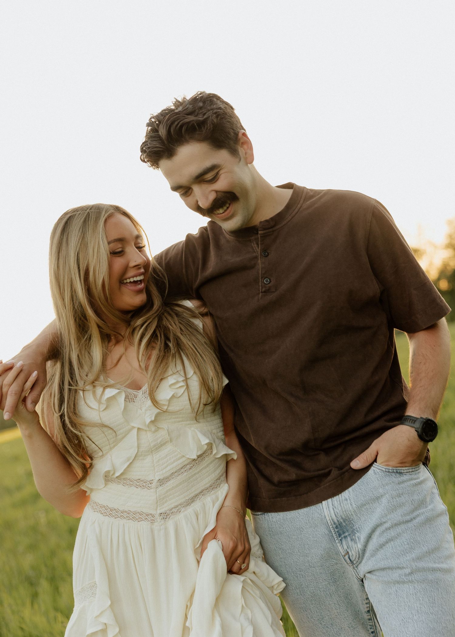 A laughing couple in golden hour light, woman in white ruffled dress and man in brown shirt embracing.