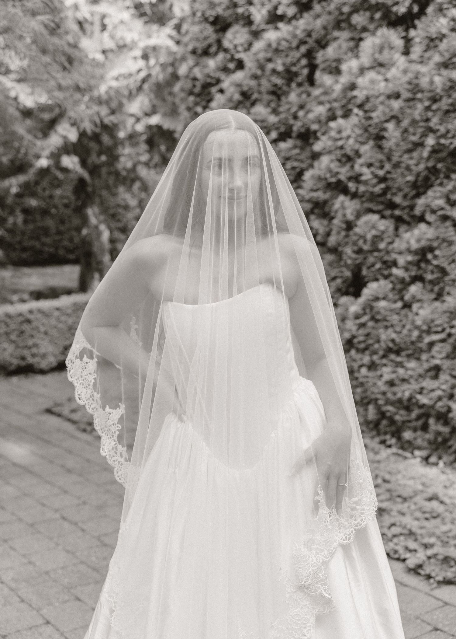 Bride in strapless gown wearing a lace-trimmed cathedral veil over her face in a black and white portrait.