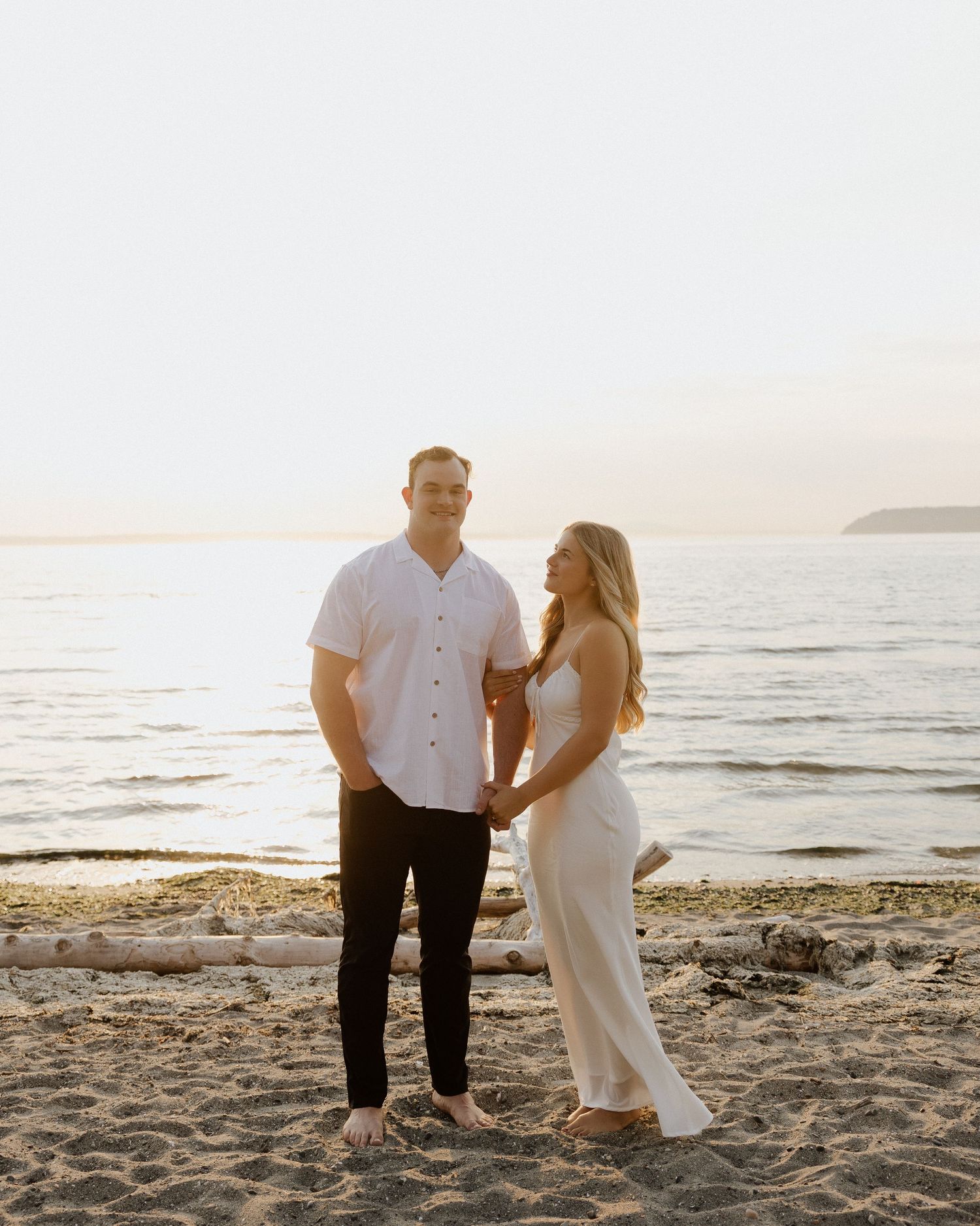 A couple stands together on a rocky beach shoreline at sunset wearing white and black.