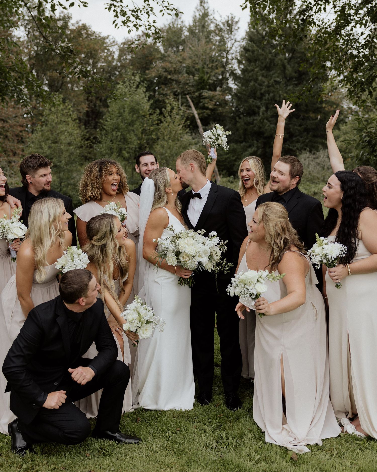 A joyful wedding party celebrates with raised arms and white bouquets on a grassy lawn.