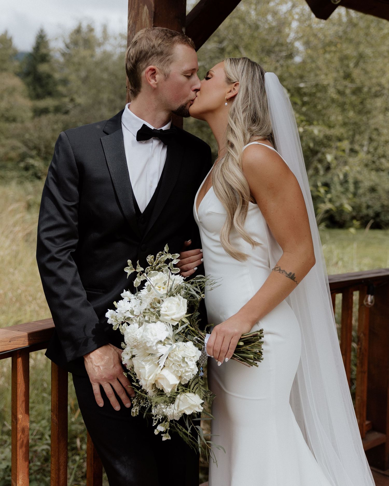 Romantic wedding couple sharing intimate moment on wooden deck with lush greenery backdrop.