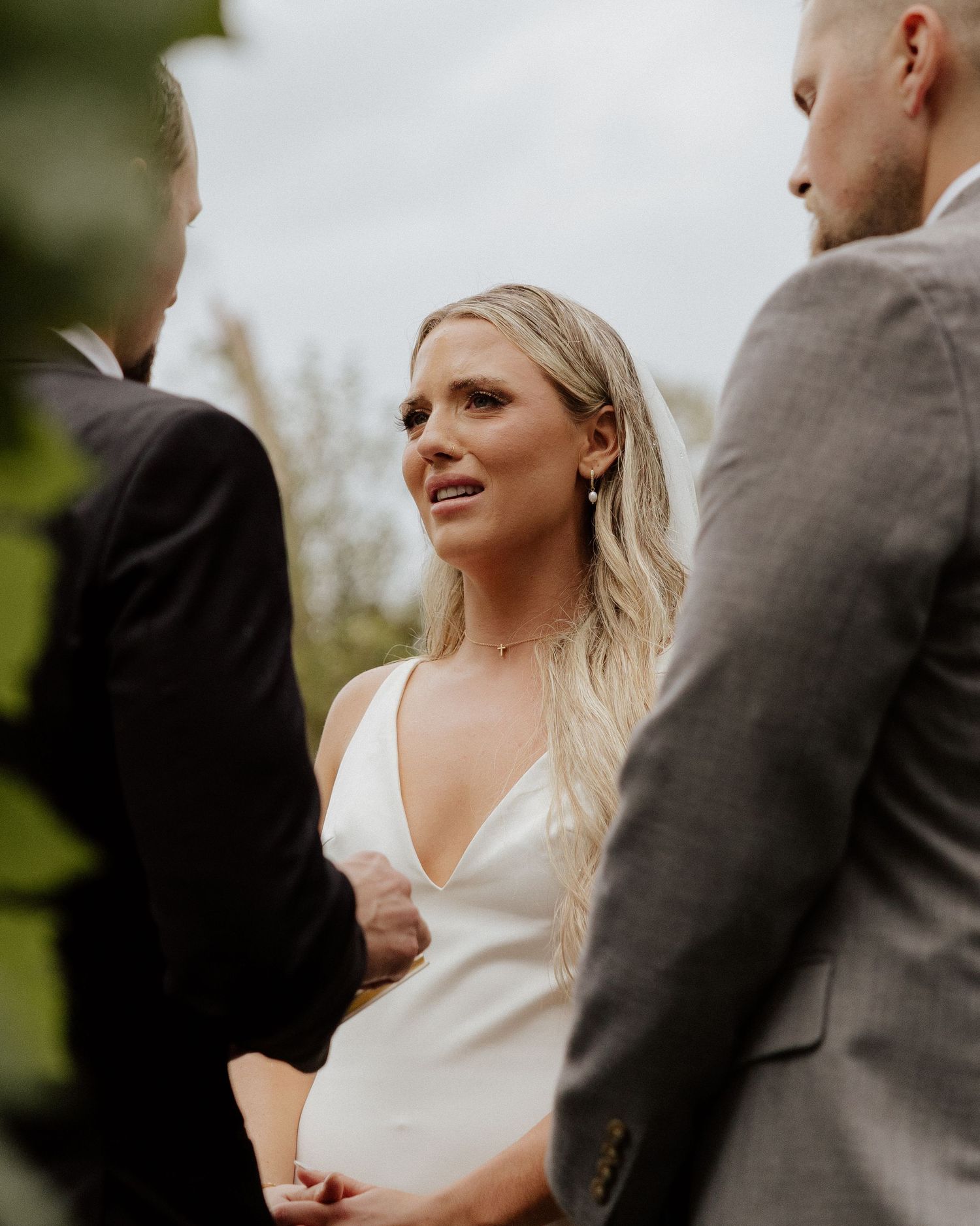 A bride in a sleek white gown listens during an outdoor wedding ceremony.