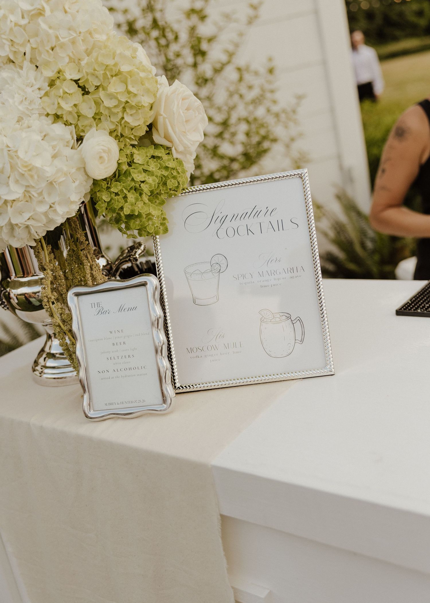 Elegant wedding signature cocktails sign in silver frame beside white hydrangea floral arrangement.