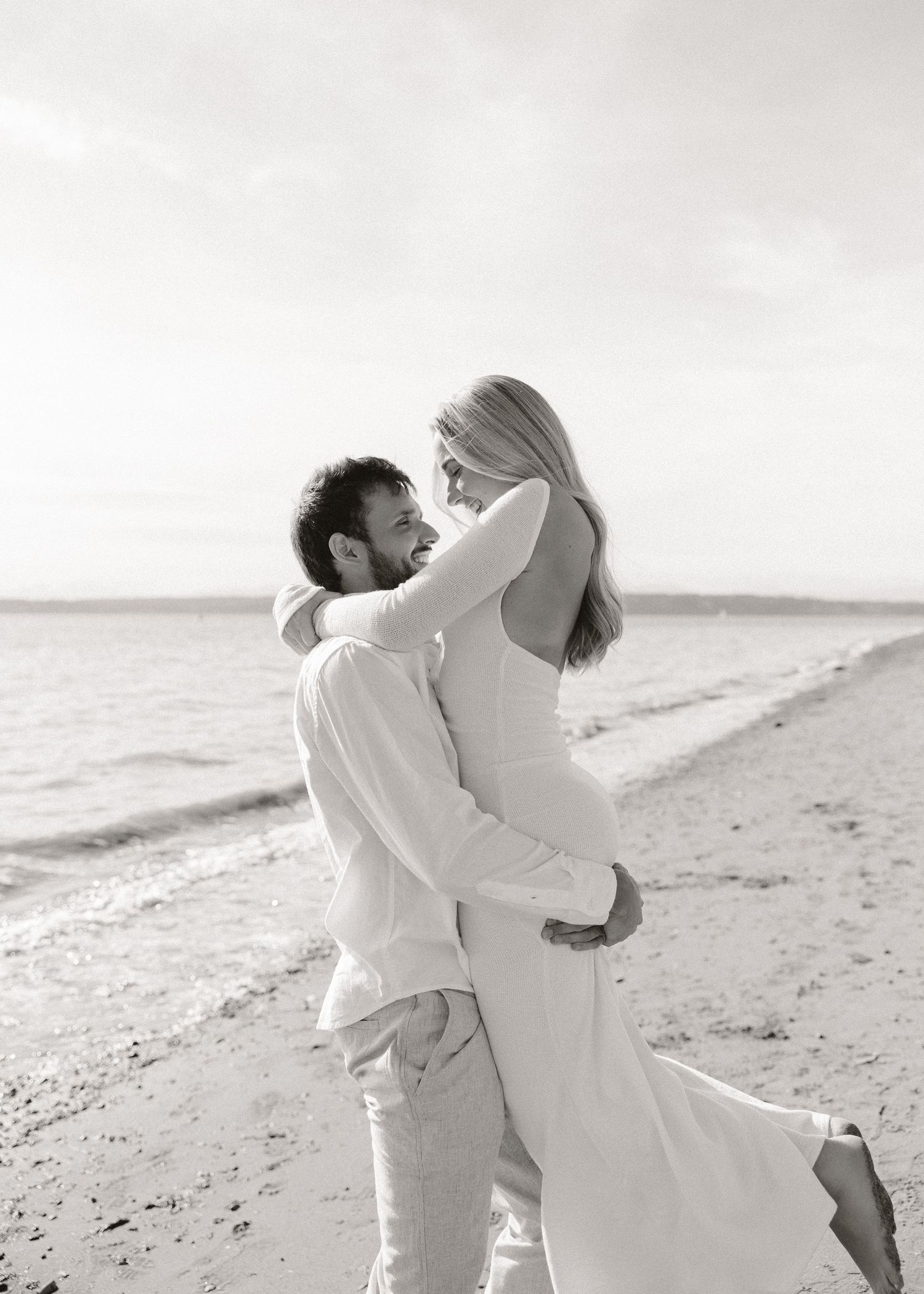 Black and white photo of a couple embracing on a beach, woman in elegant backless gown, man in white shirt.